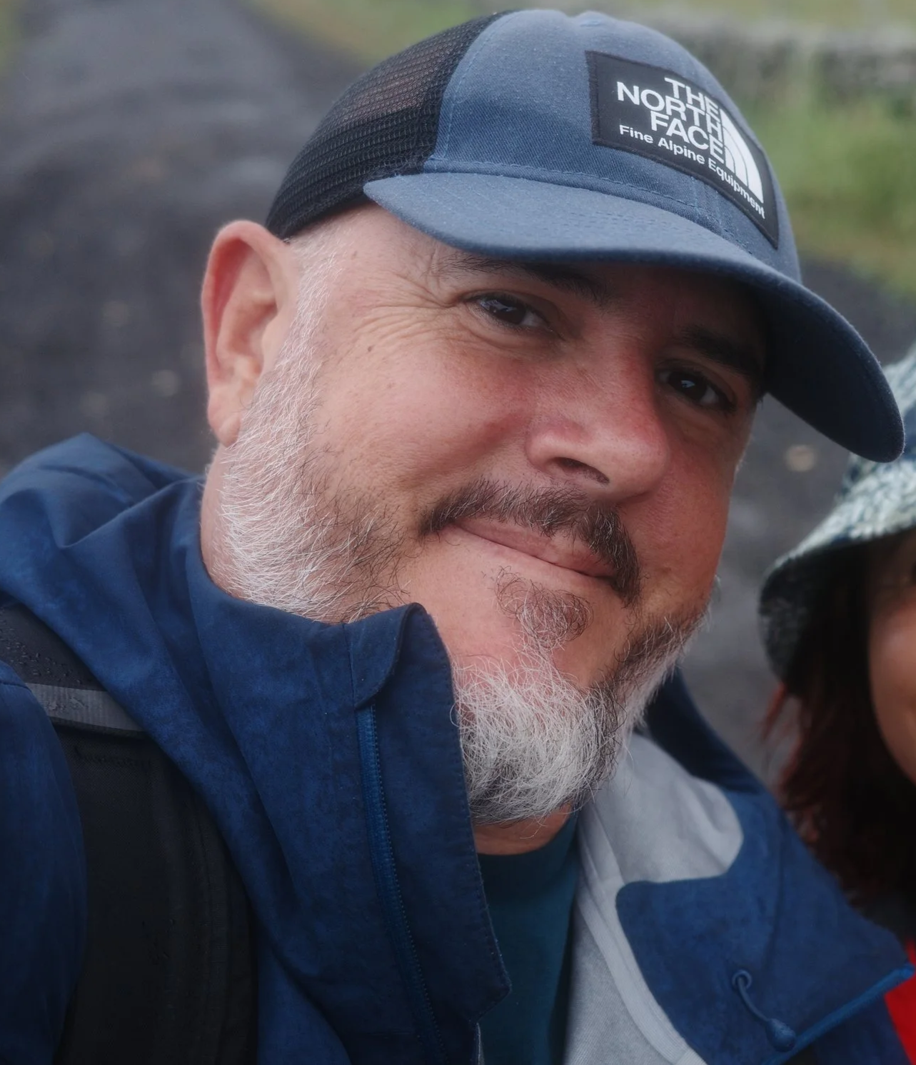A close-up of a man with a gray beard and mustache wearing a blue and black North Face cap and a blue jacket, smiling outdoors.