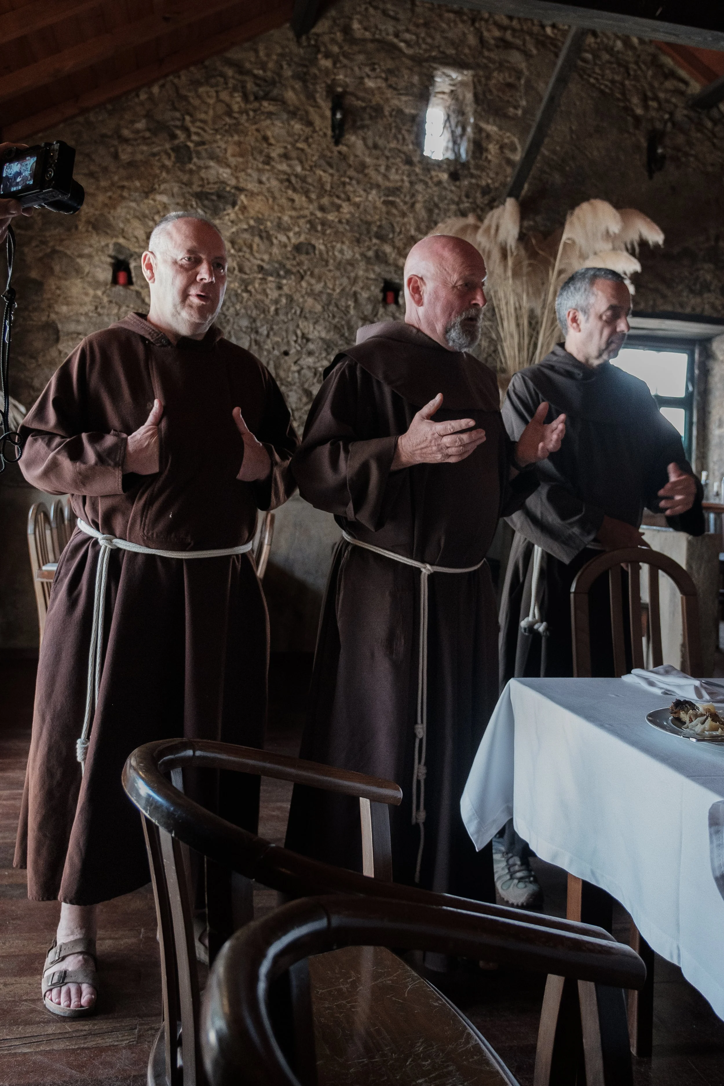 Three men dressed in brown robes with ropes at their waists, standing in a rustic room with exposed stone walls, possibly during a religious or cultural ceremony. One man on the left is making a gesture with his hands, and a camera is visible on the 