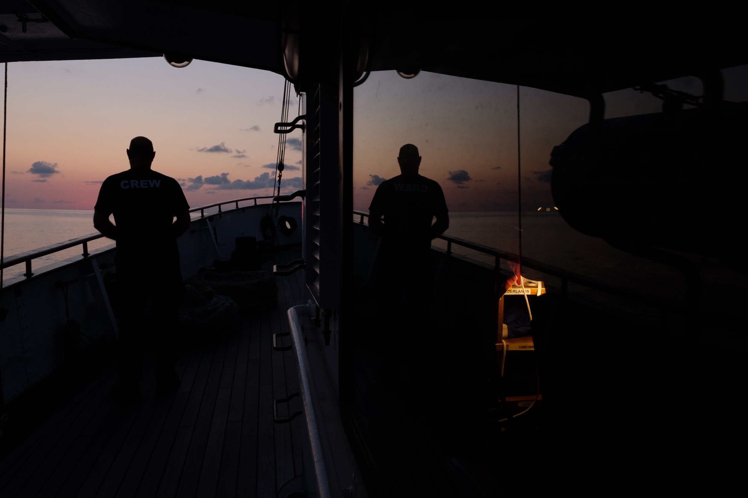 Silhouettes of crew members on a ship's deck at sunset, with a sunset sky and ocean in the background.