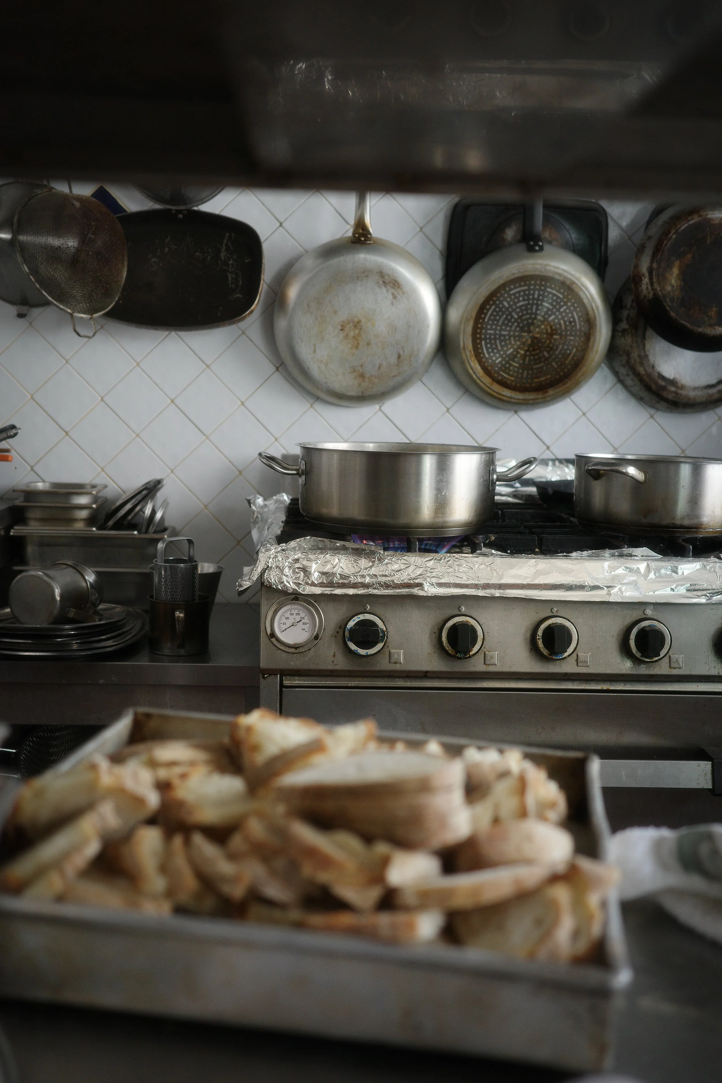 Commercial kitchen with cooking pots on stove, hanging saucepans and strainers on the wall, and a tray of sandwiches in the foreground.