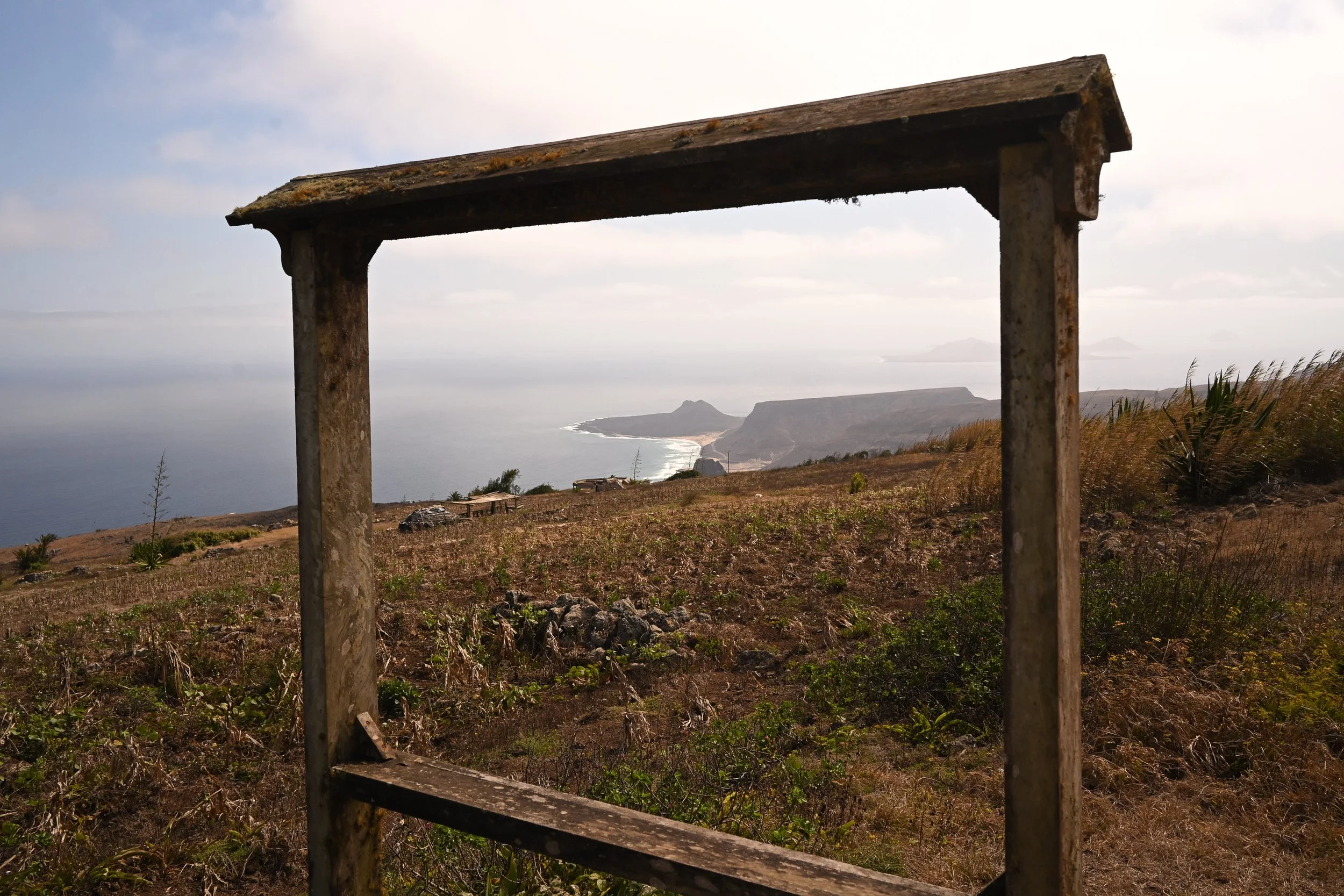 An old, weathered wooden frame standing on a hill overlooking a coastline with cliffs and the ocean. The sky is partly cloudy.