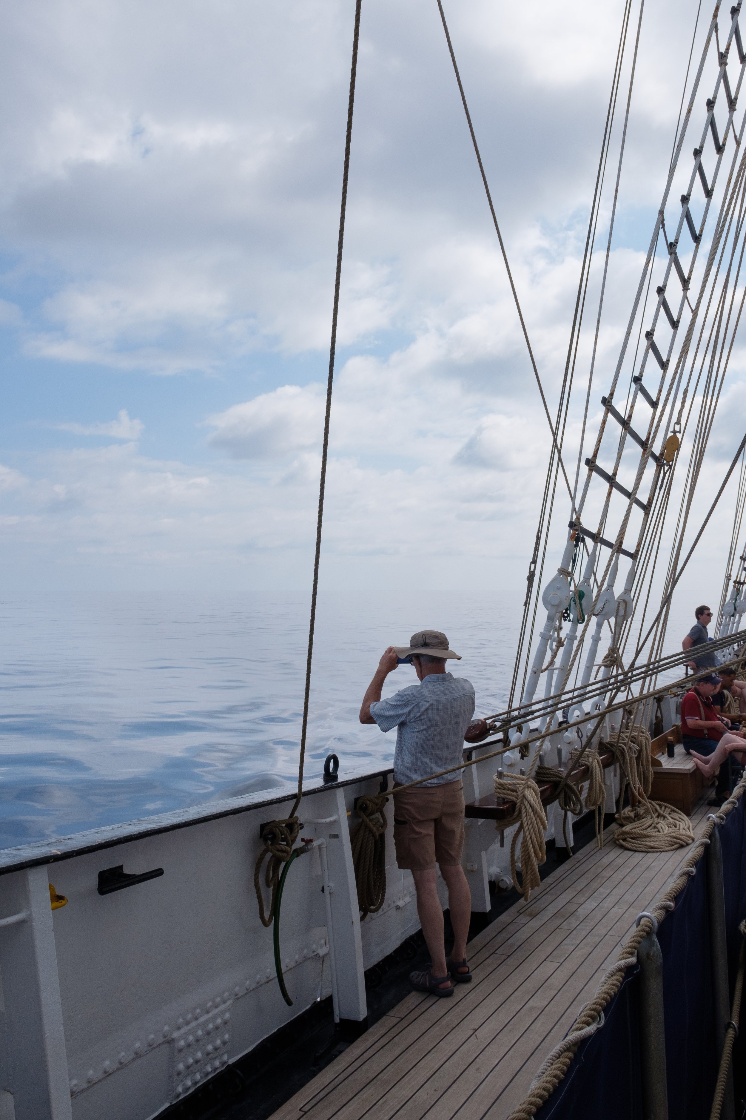 People on a ship's deck with ropes and rigging, under a cloudy sky, some individuals looking out at the water, one person in a hat taking a photo.