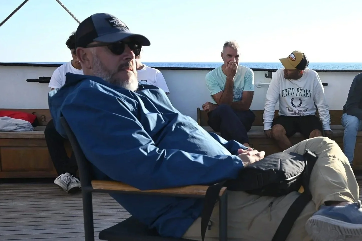 Group of men sitting and talking on a boat deck with ocean in the background, some wearing casual clothing and hats.