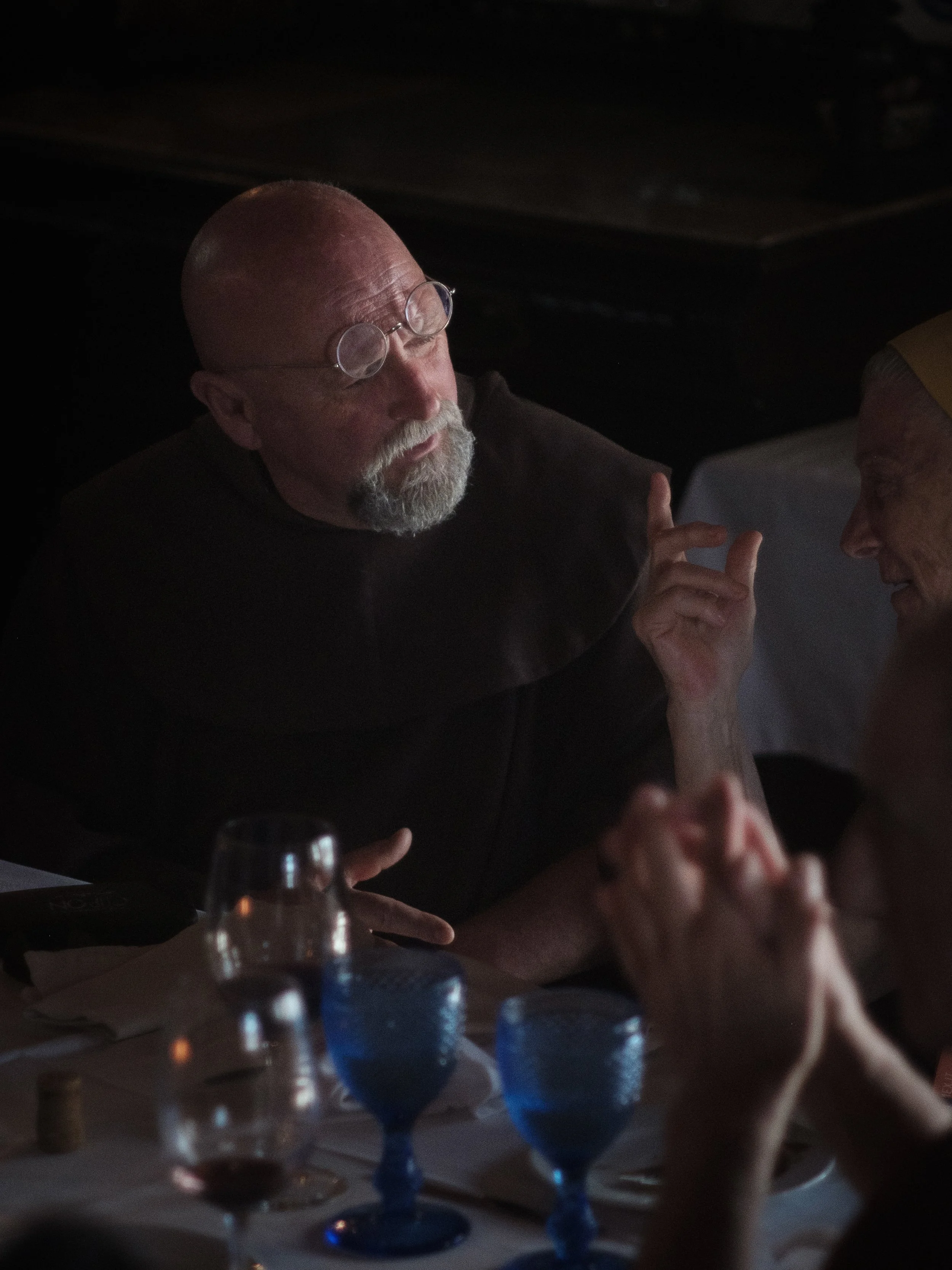 A man with glasses and a beard talking to an older woman at a table with bottles and glasses in a dimly lit setting.