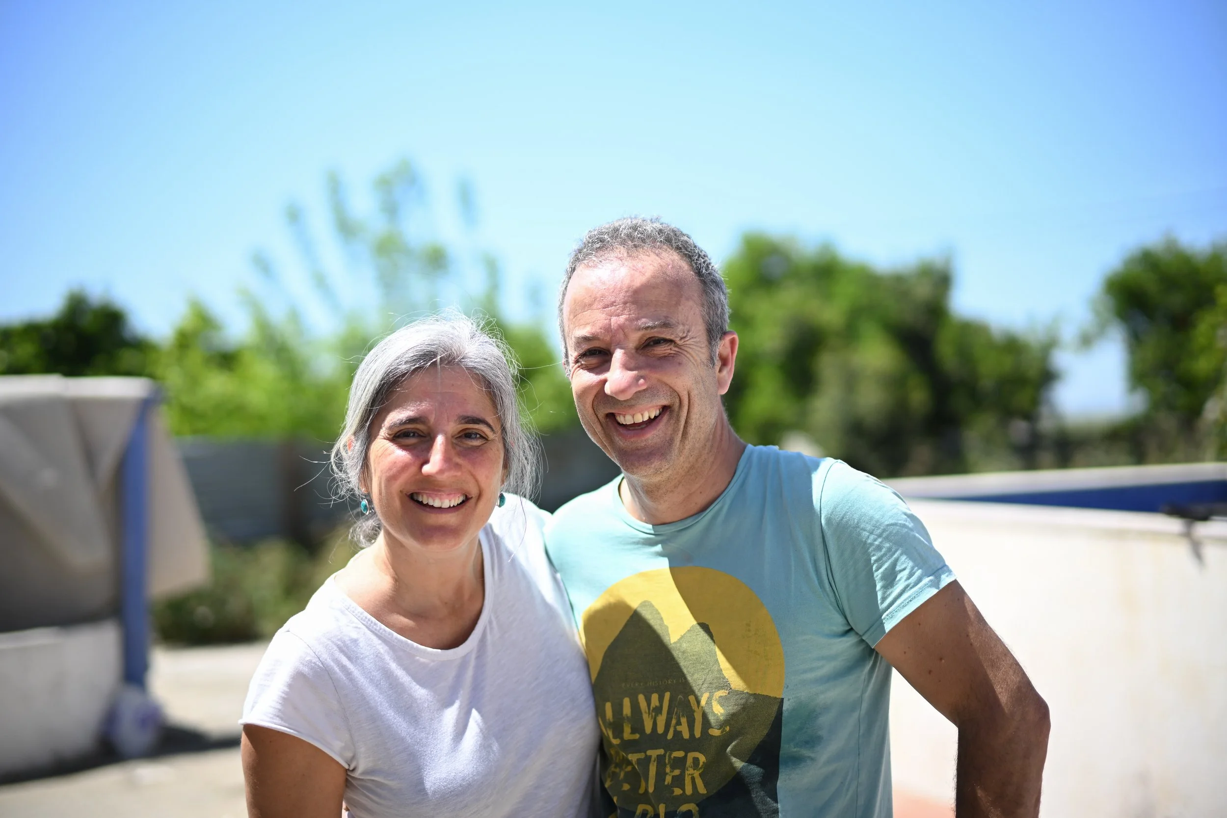 A smiling older woman with gray hair and a younger man with short gray hair taking a selfie outdoors on a sunny day, with green trees and blue sky in the background.