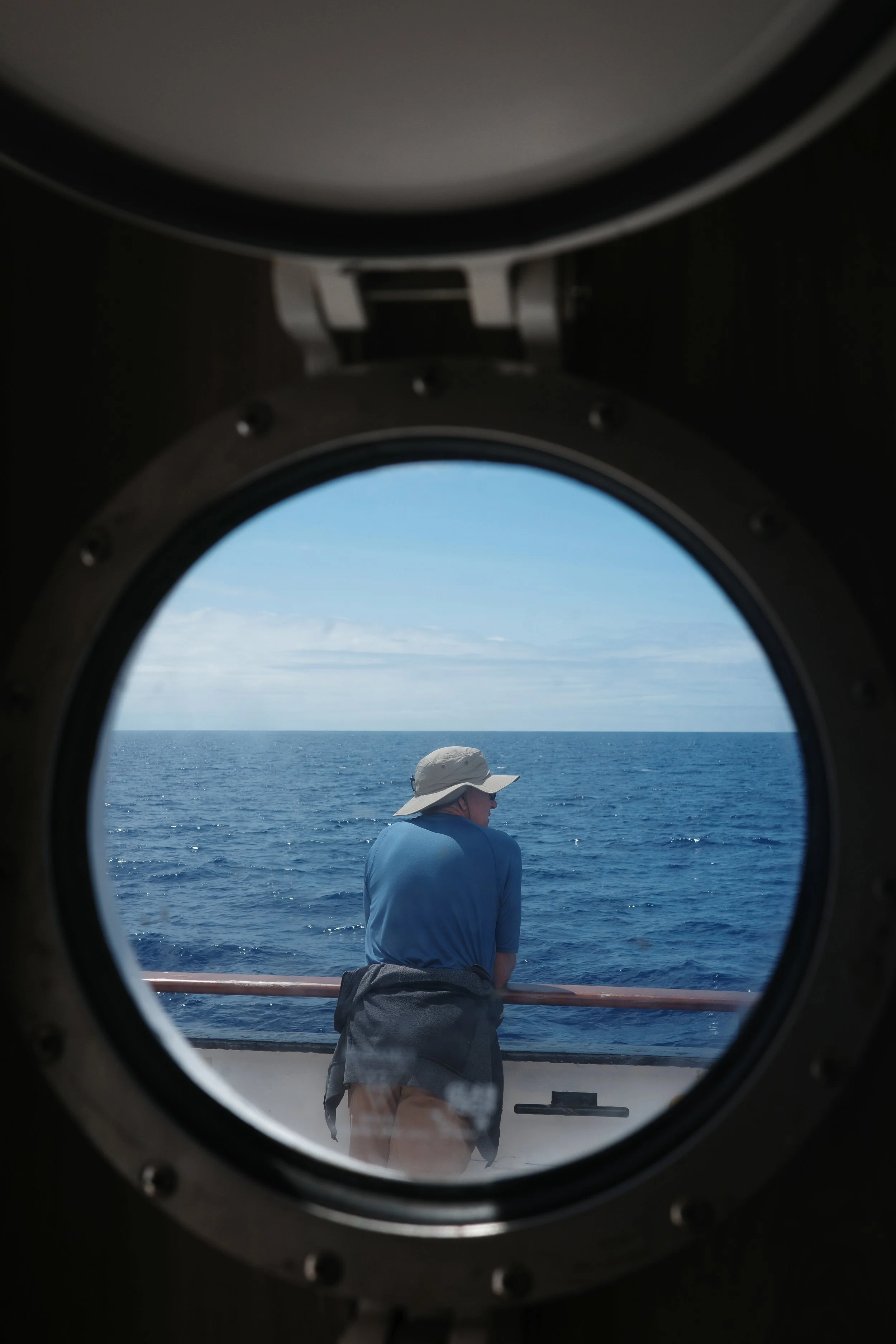 View of the ocean through a porthole, with a man wearing a hat and blue jacket standing on a ship's deck, looking at the sea.