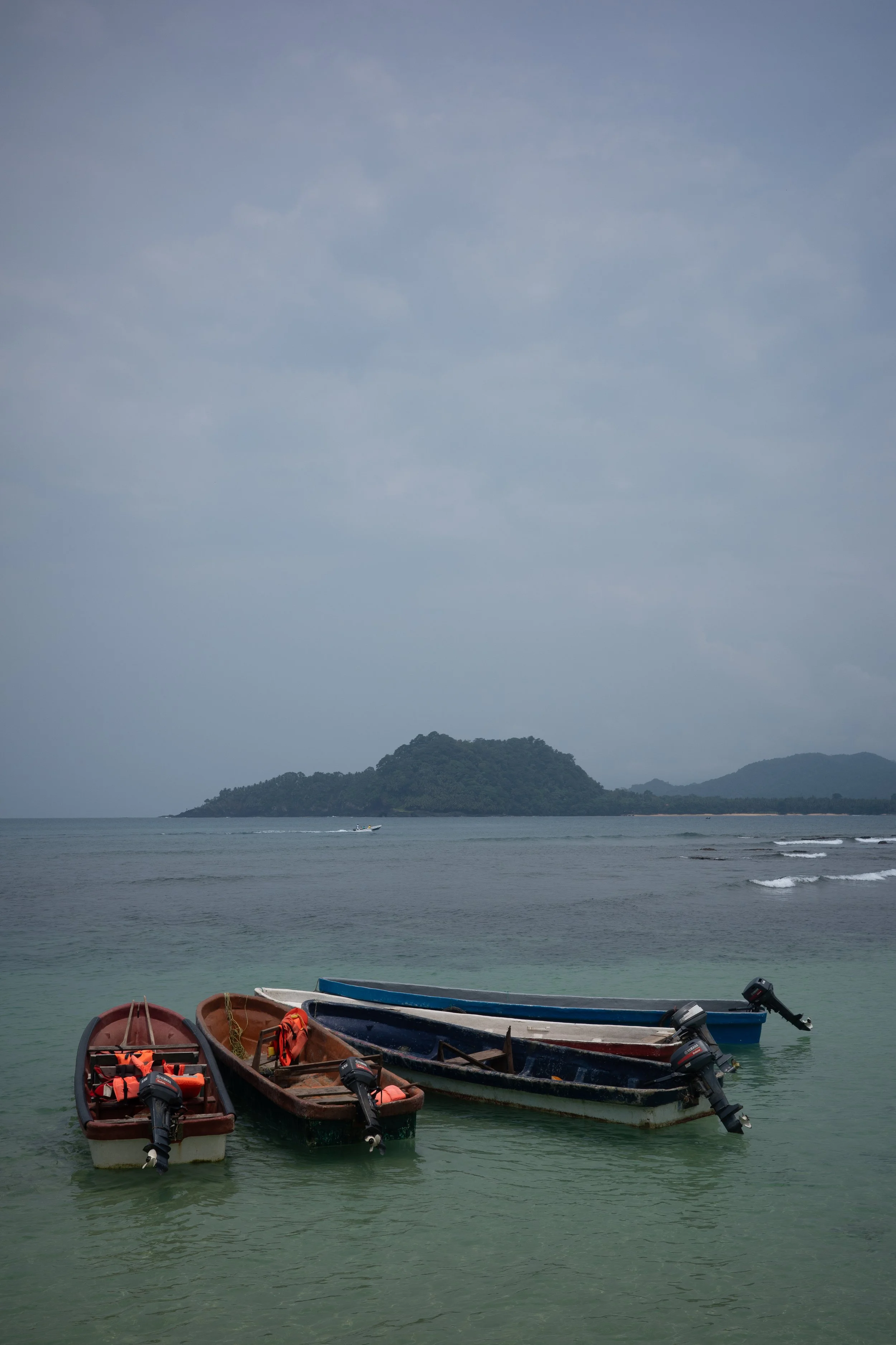 Boats anchored near the shore with hills and cloudy sky in the background, and a small boat speeding across the water.