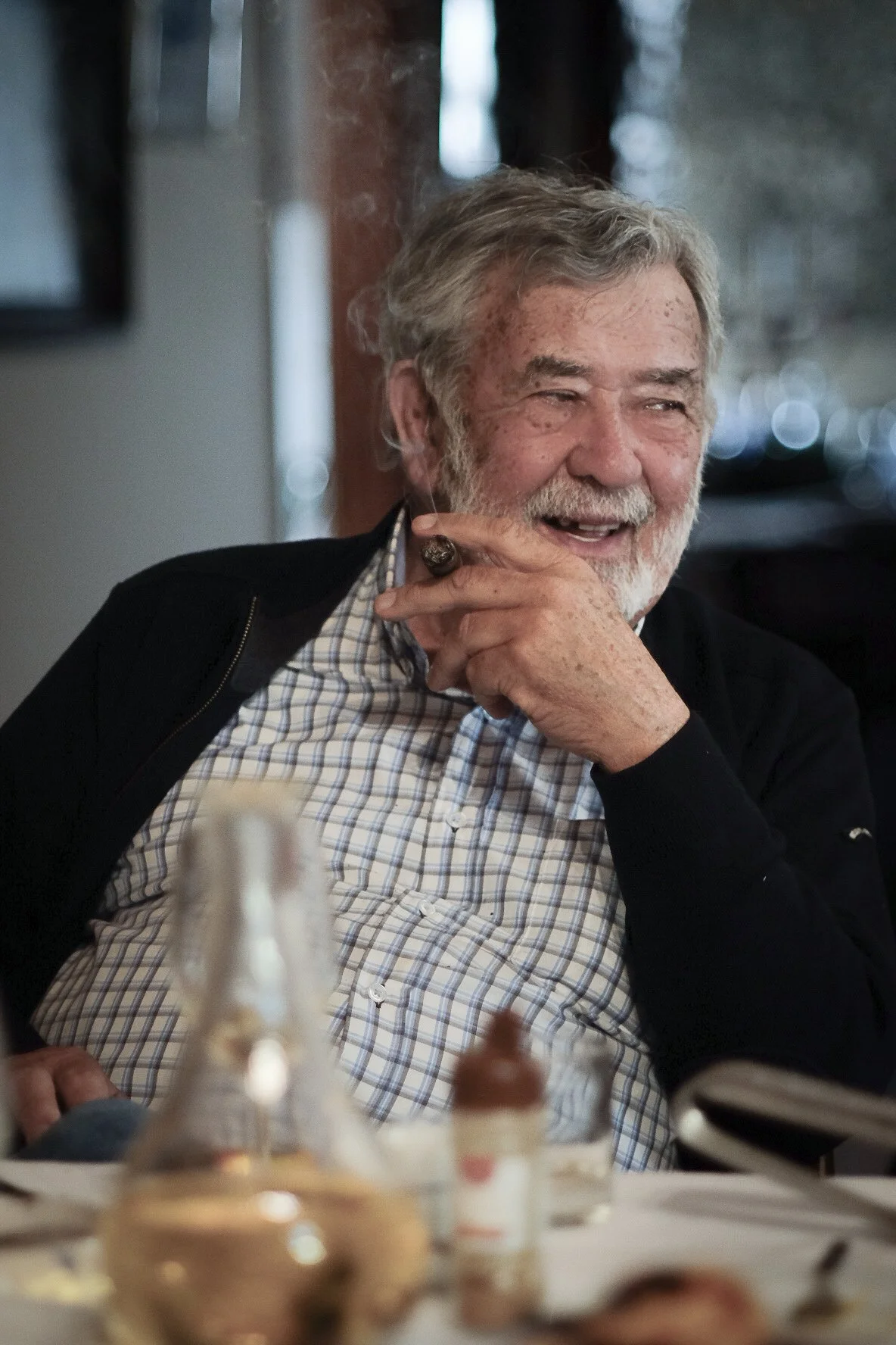 An older man with gray hair and a beard, smiling and laughing, sitting at a table with cake and desserts in front of him.
