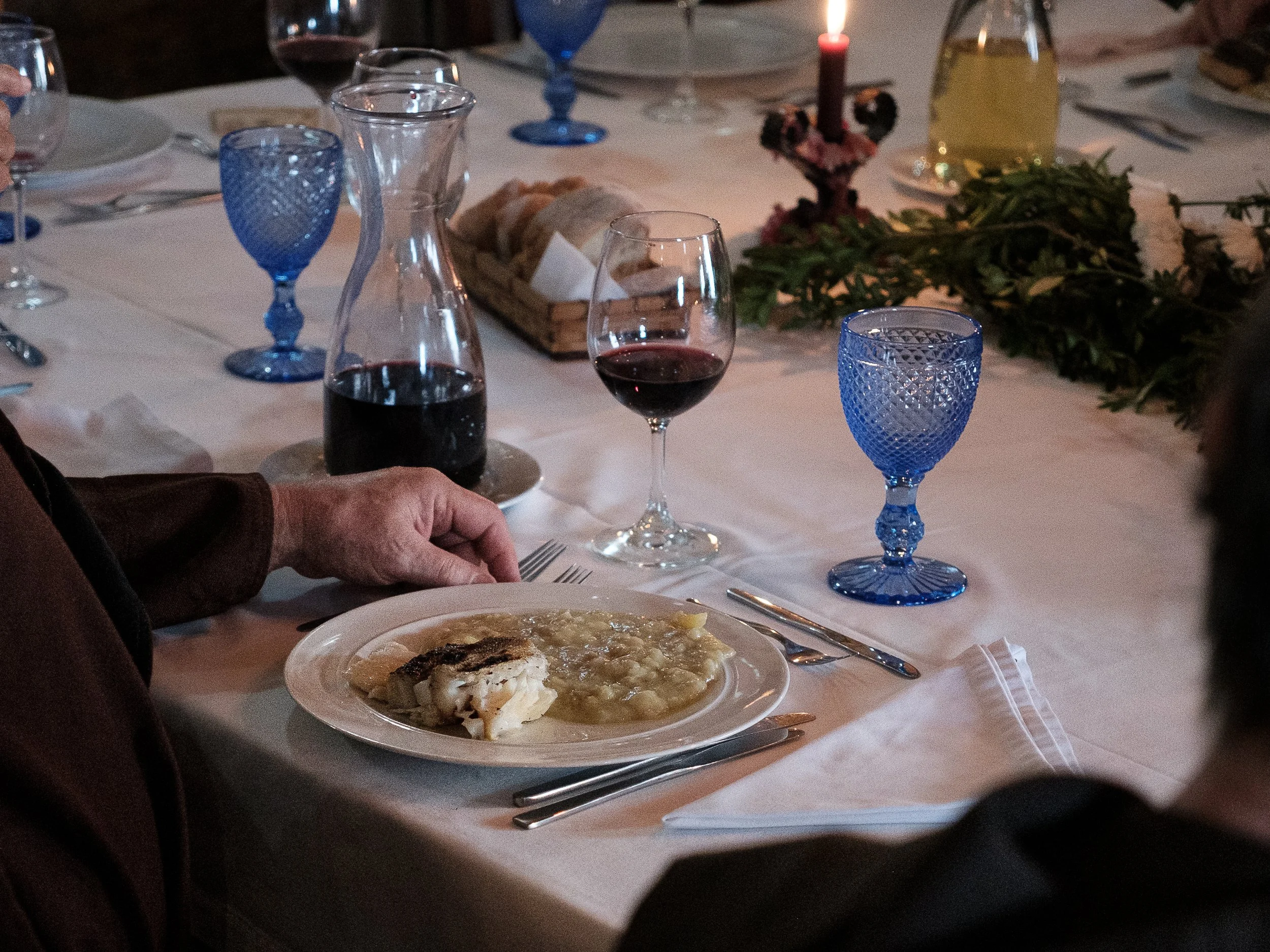 A dinner table set with blue glassware, wine glasses filled with red wine, a lit candle, a basket of bread, and a plate of food, with some greenery as a centerpiece.