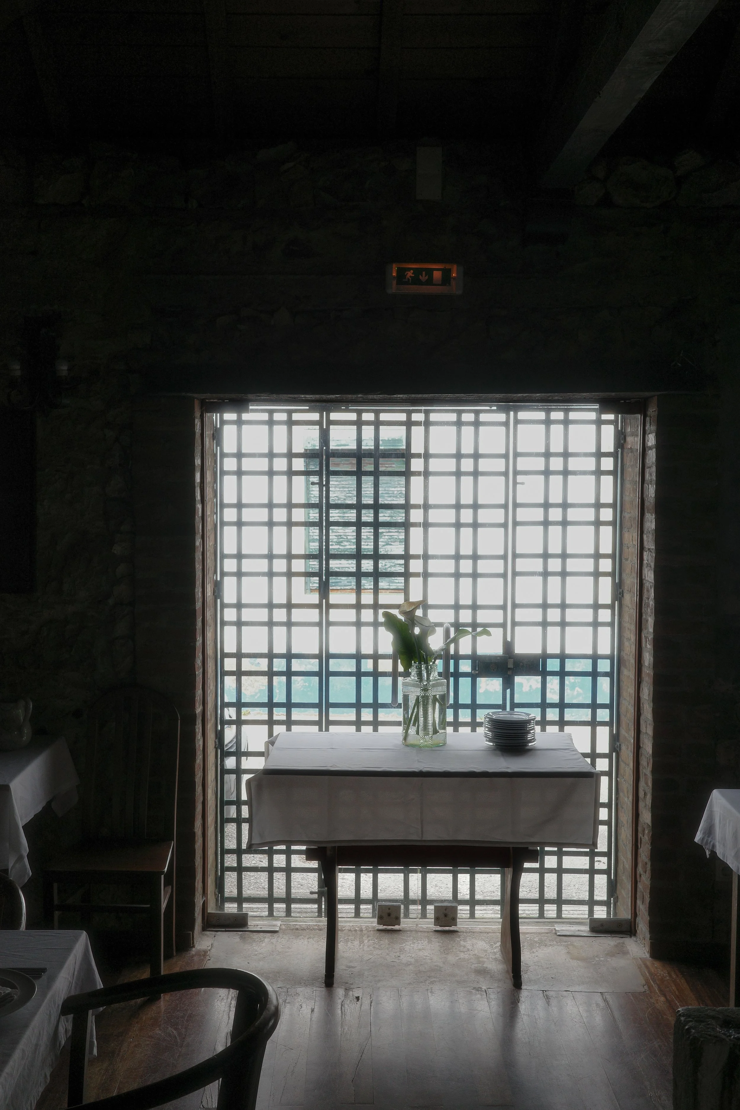 A dining area with a table covered with a white tablecloth, a glass jar with a flower, and a stack of plates placed on the table. The table is positioned in front of a large window with a grid pattern, allowing natural light to brighten the room.