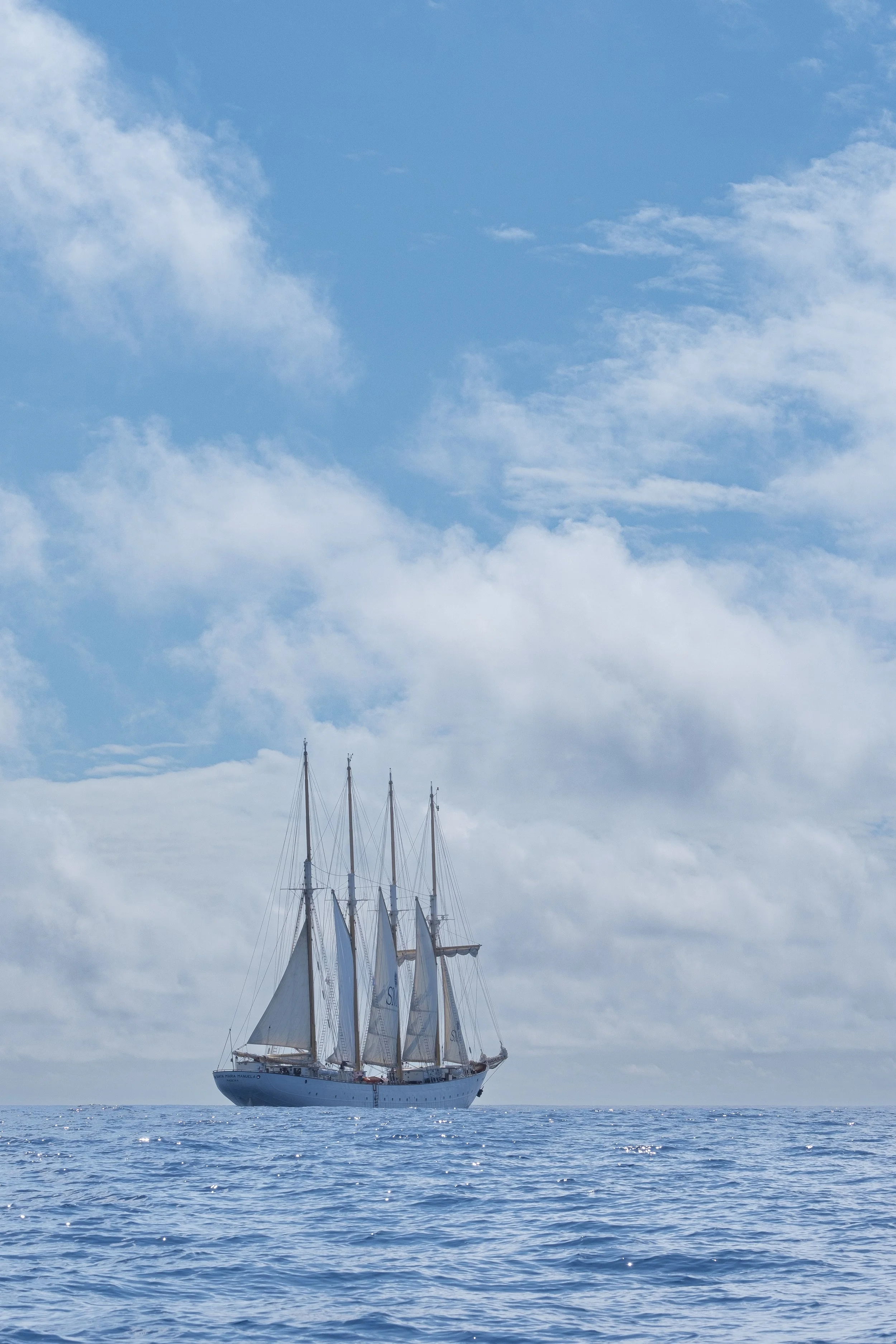 A large sailing ship with multiple masts and sails on calm ocean waters under a partly cloudy sky.
