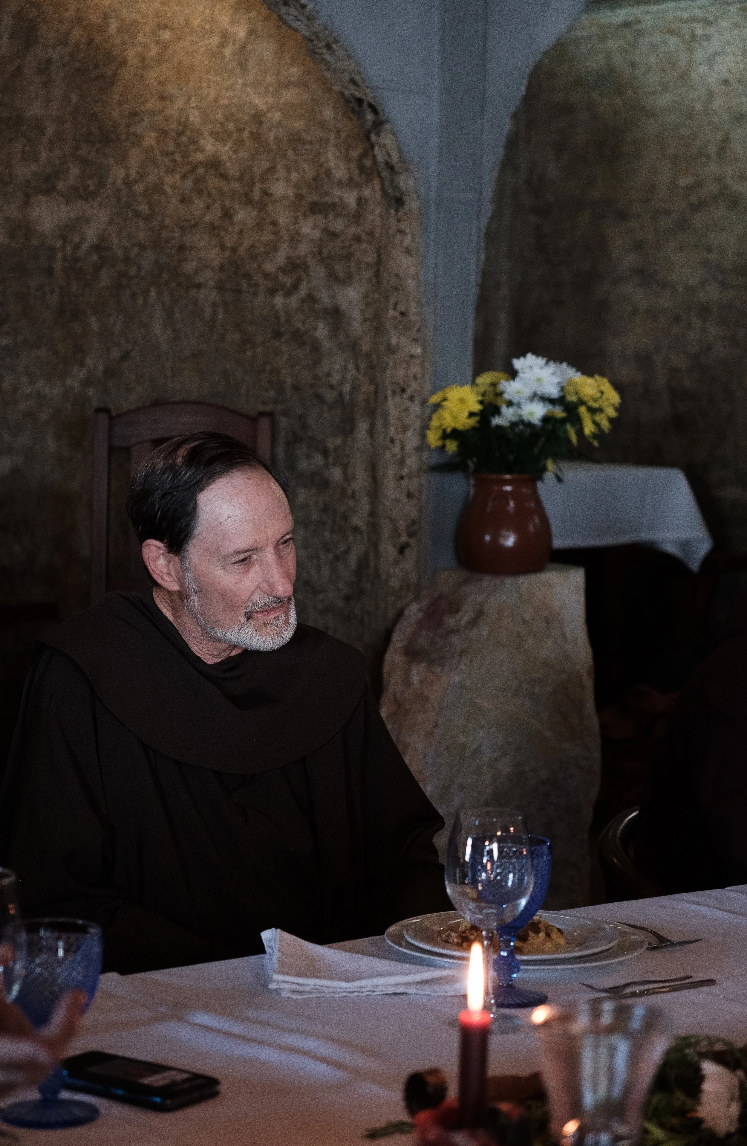 A man with dark hair and a beard sitting at a table with a plate of food in front of him. The table has a white tablecloth, glasses, and utensils. There is a candle in the foreground and a vase with yellow and white flowers in the background, set aga