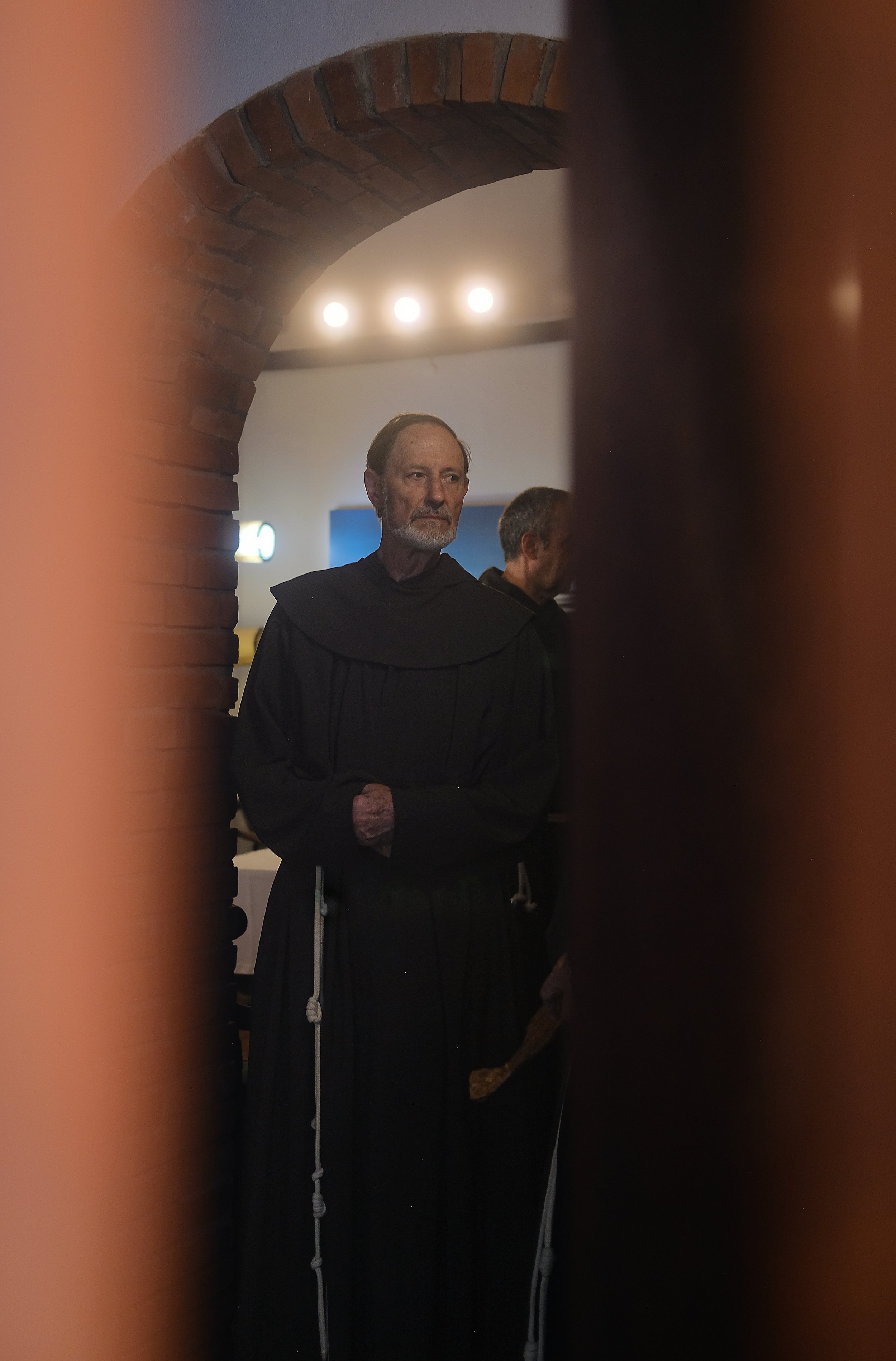 A priest or monk in black robes standing behind a partially open door or window with a brick arch and soft lighting in the background.