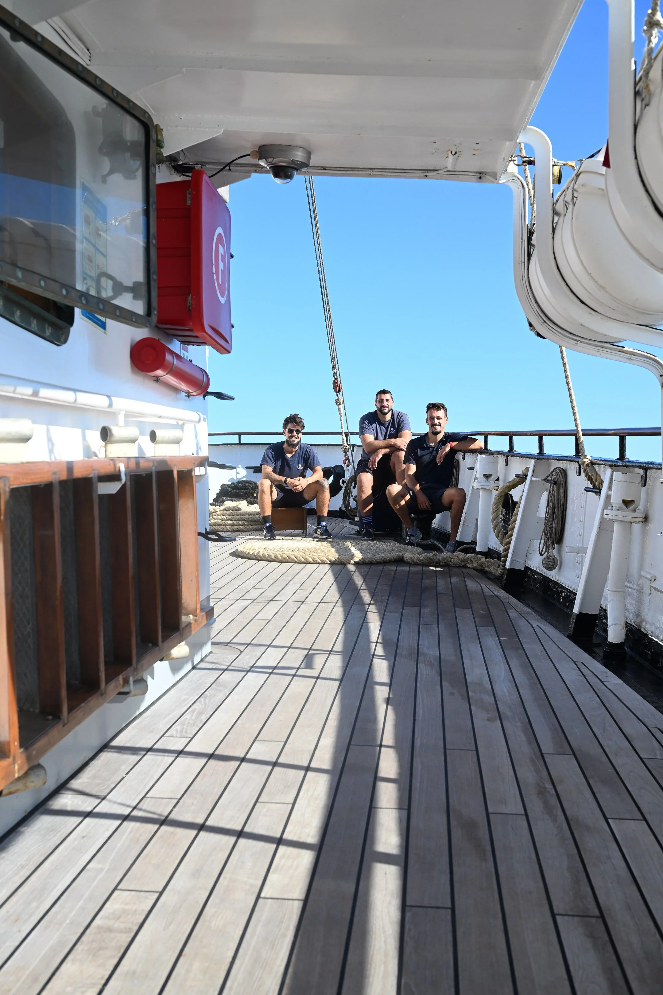 Three men sitting on the deck of a ship, smiling. The deck has wooden planks, and there are ropes and equipment around them. The sky is clear and blue.