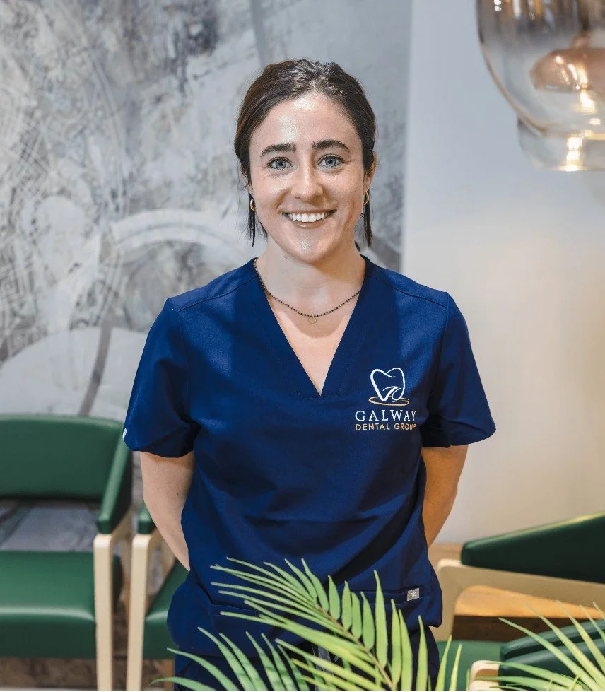 A young woman with brown hair, blue eyes, and a friendly smile, wearing navy scrubs with the logo 'Galway Dental Group,' standing in a modern dental office with a textured wall, green chairs, a hanging light fixture, and a potted plant in front of her.