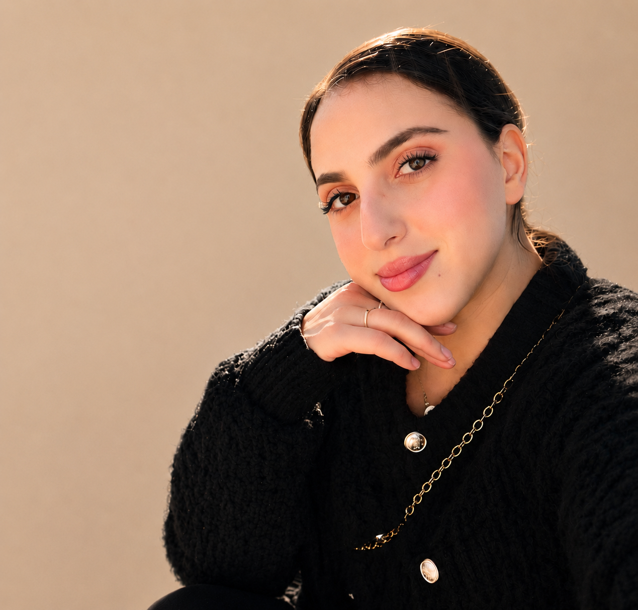 A woman with dark hair and brown eyes smiling, wearing a black textured sweater, a gold necklace, and a gold ring, with her hand resting near her chin, illuminated by soft sunlight.