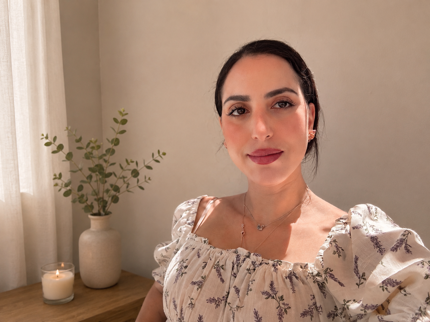 A woman taking a selfie in a softly lit room. She is wearing a light-colored, floral-patterned puff-sleeve dress and jewelry, with a neutral expression. In the background, there is a potted plant, a lit candle, and a wooden table, with cream-colored curtains on the side.