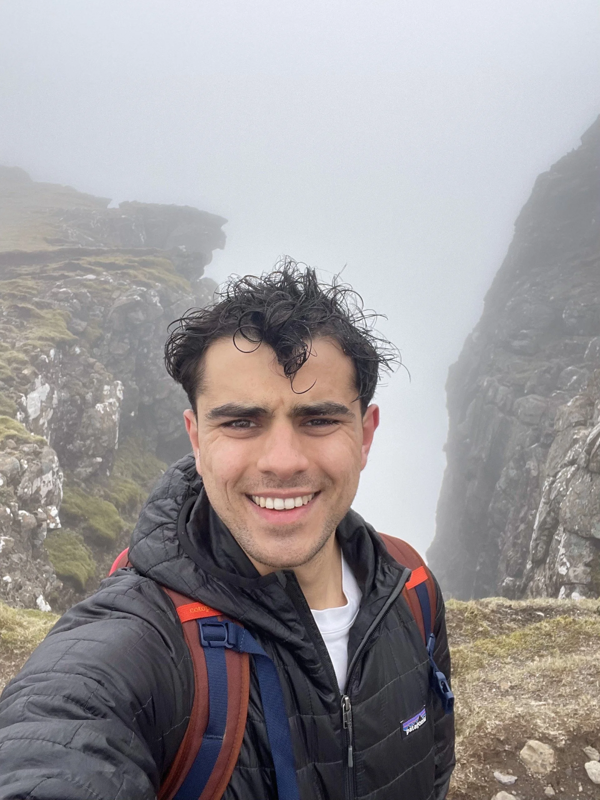 Dom Stoddart smiling for a selfie on a hiking trail between tall, rocky mountains with moss and patches of grass, surrounded by fog.