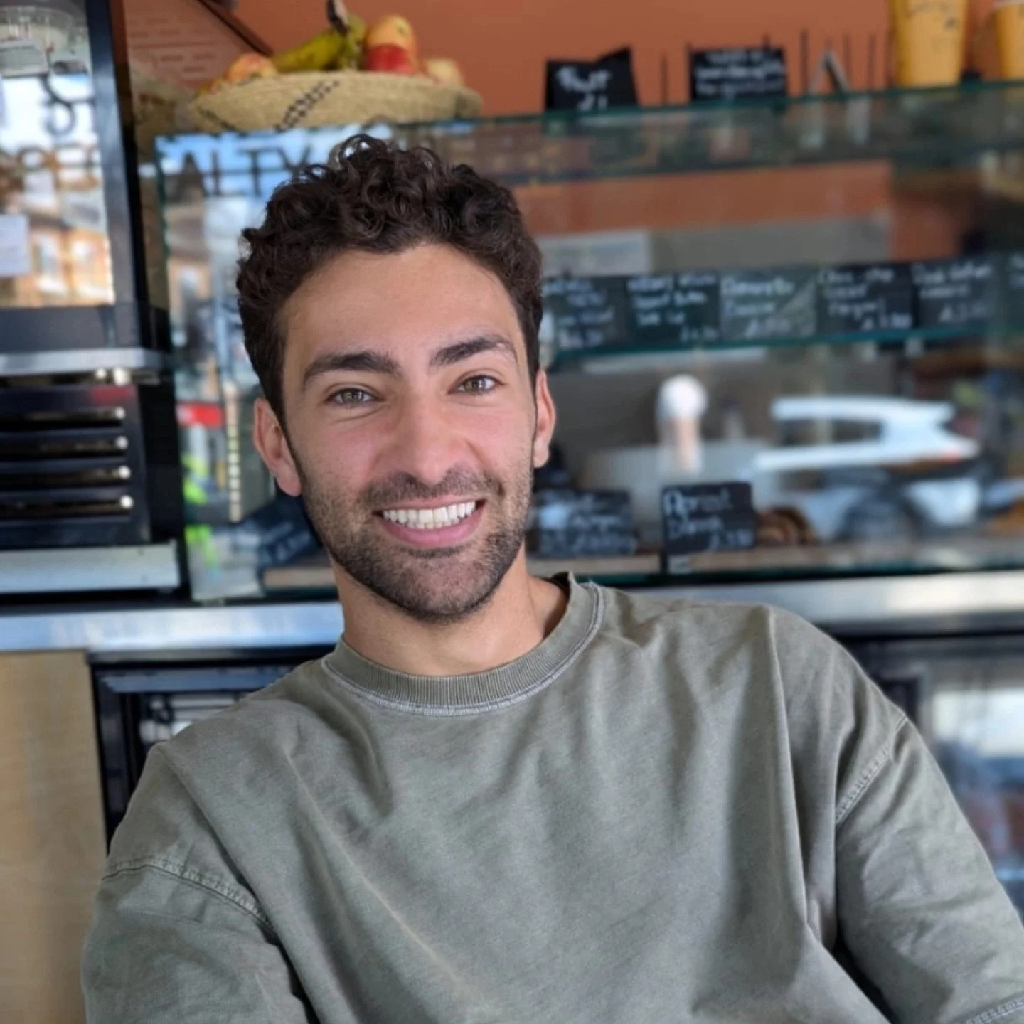 Imre Gerrits smiling, sitting inside a cafe with a glass display case behind him containing baked goods and chalkboard menus.