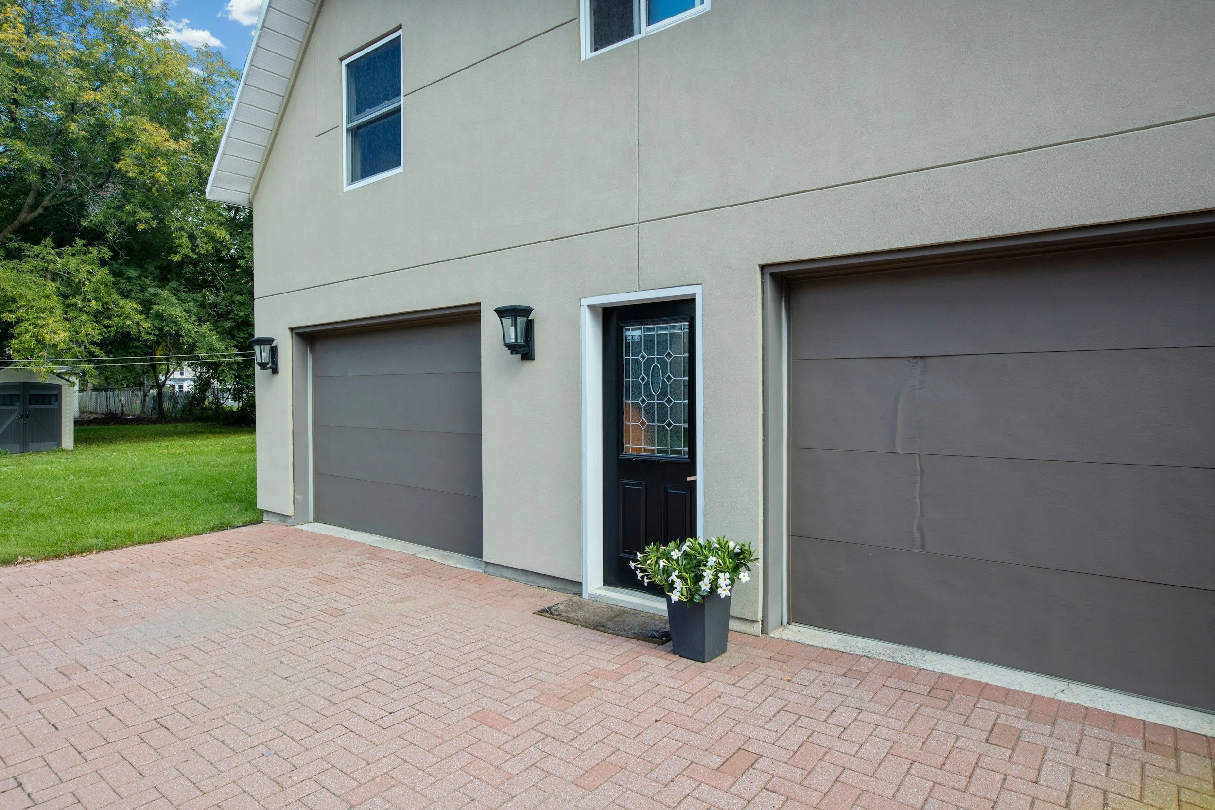 Exterior view of a modern residential building with two garage doors, a decorative front door with a black finish, and two black lantern-style wall lights. There is a potted plant with white flowers near the front door and a brick driveway in front of the garages. Green lawn and trees are visible in the background under a partly cloudy sky.