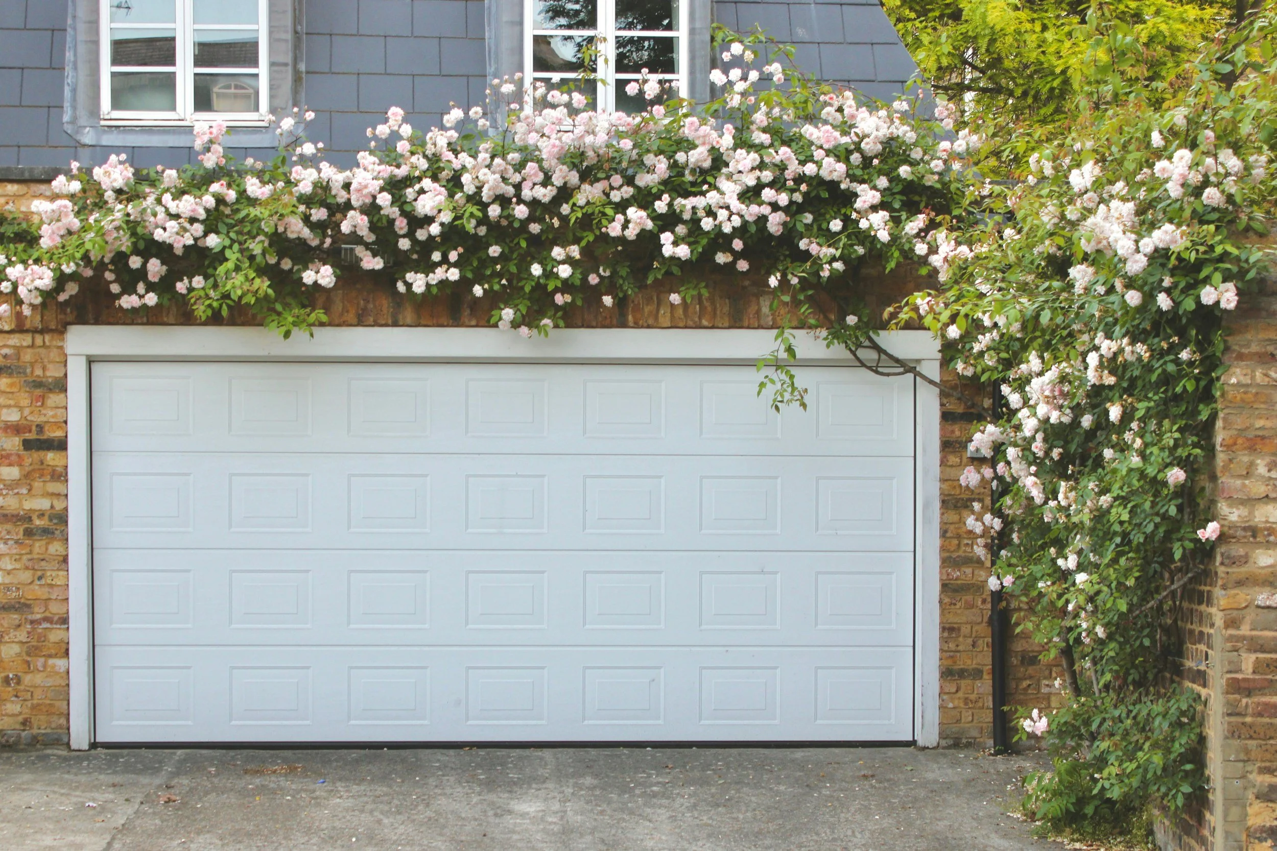 A white garage door with a brick and gray shingle house exterior above it, decorated with pink and white flowering plants climbing along the brick wall and surrounding a window.