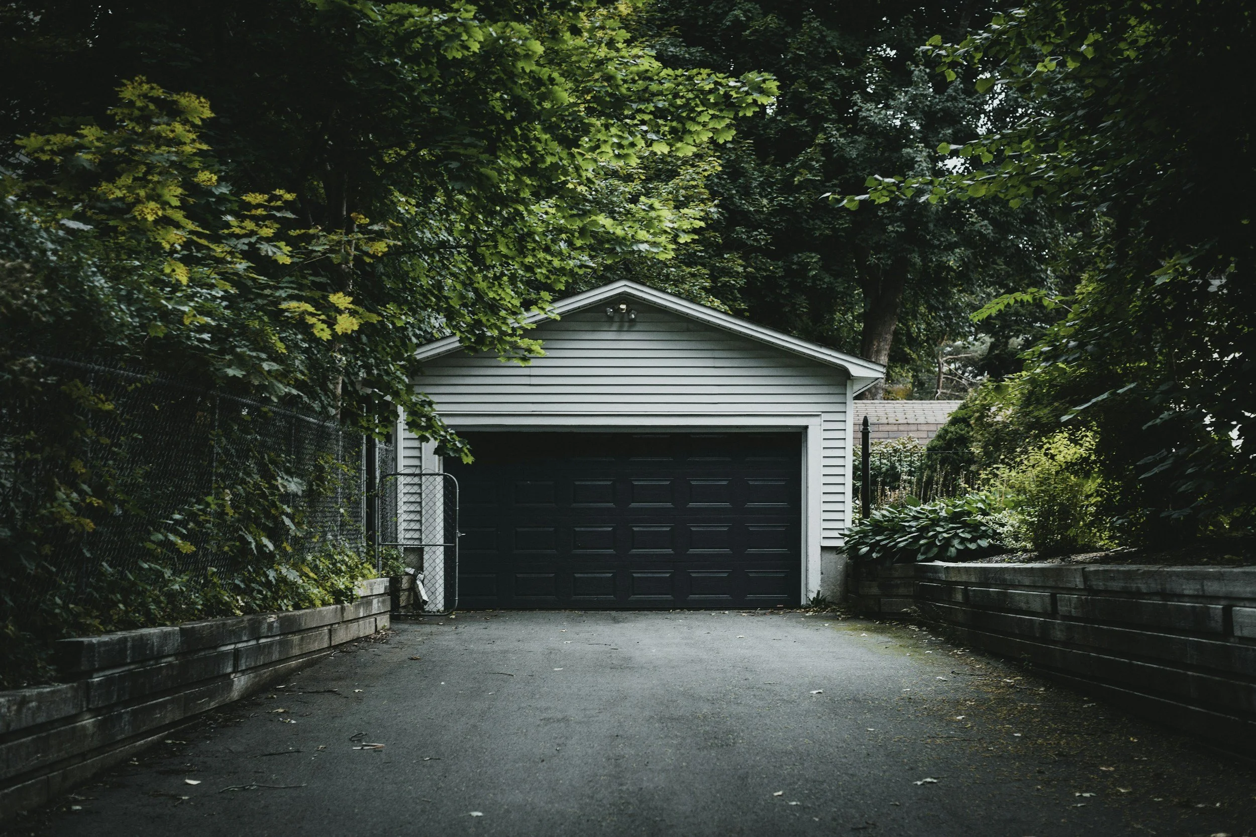 A garage with a closed dark door, surrounded by lush green trees and bushes, with a driveway leading up to it.