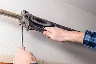 Close-up of a person in a blue plaid shirt using a hand tool to stretch a mechanical spring or cable between two hooks.