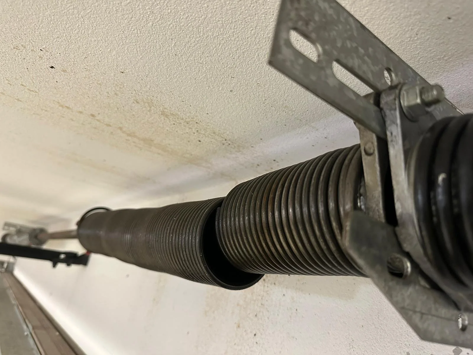 A close-up view of a garage door torsion spring assembly mounted on a ceiling, consisting of a long metal coil spring, a metal bracket, and a tension rod.