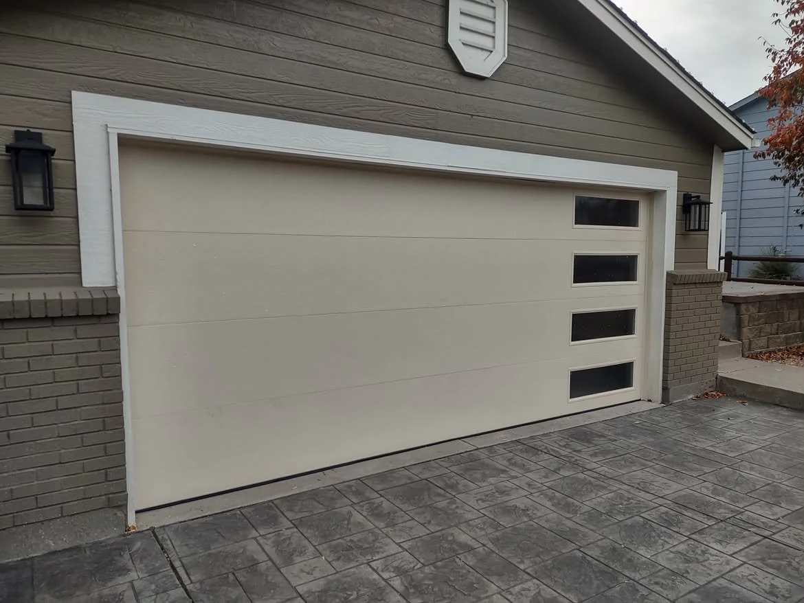 Modern garage door with horizontal panels and four small windows on the right side, attached to a house with gray siding and brick accents, flanked by two black wall-mounted lantern lights.