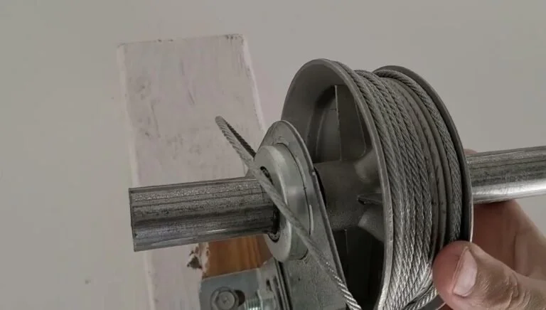 Close-up of a person’s hand holding a metal pulley with a steel cable wound around it, mounted on a vertical surface with a white wall in the background.