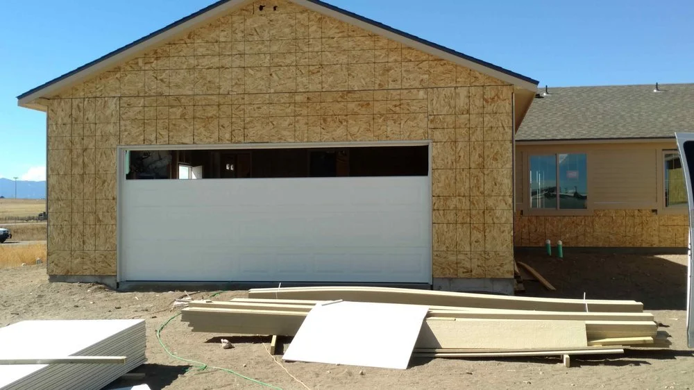 A house under construction with exterior walls covered in plywood sheathing. There is a white garage door and a side window. Building materials are stacked in front on the ground.