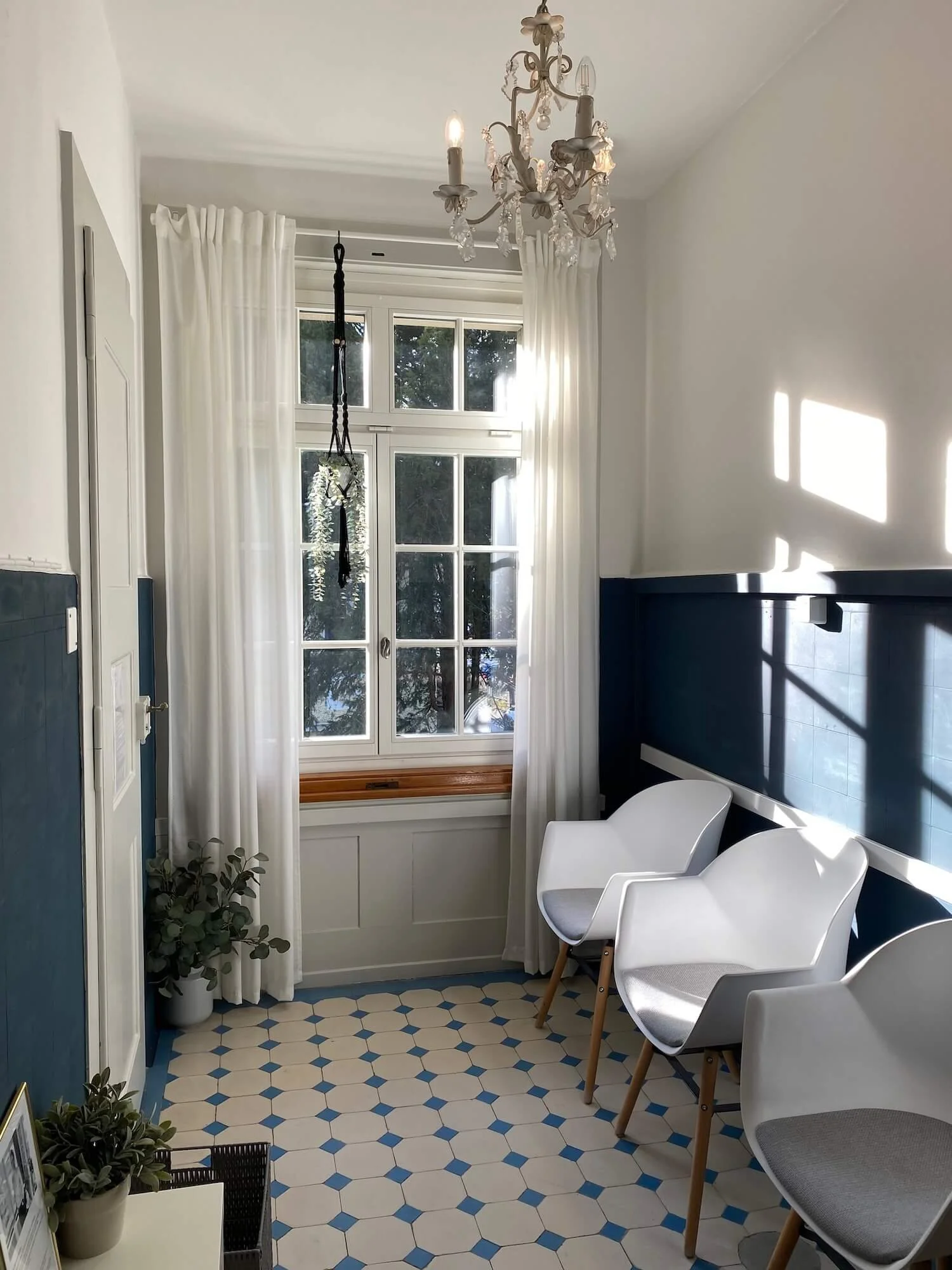 Ein heller Raum mit einem Fenster, weißen Vorhängen und Stühlen. Es gibt eine Kronleuchterdecke und einen Fliesenboden mit blau-weißen Mustern.