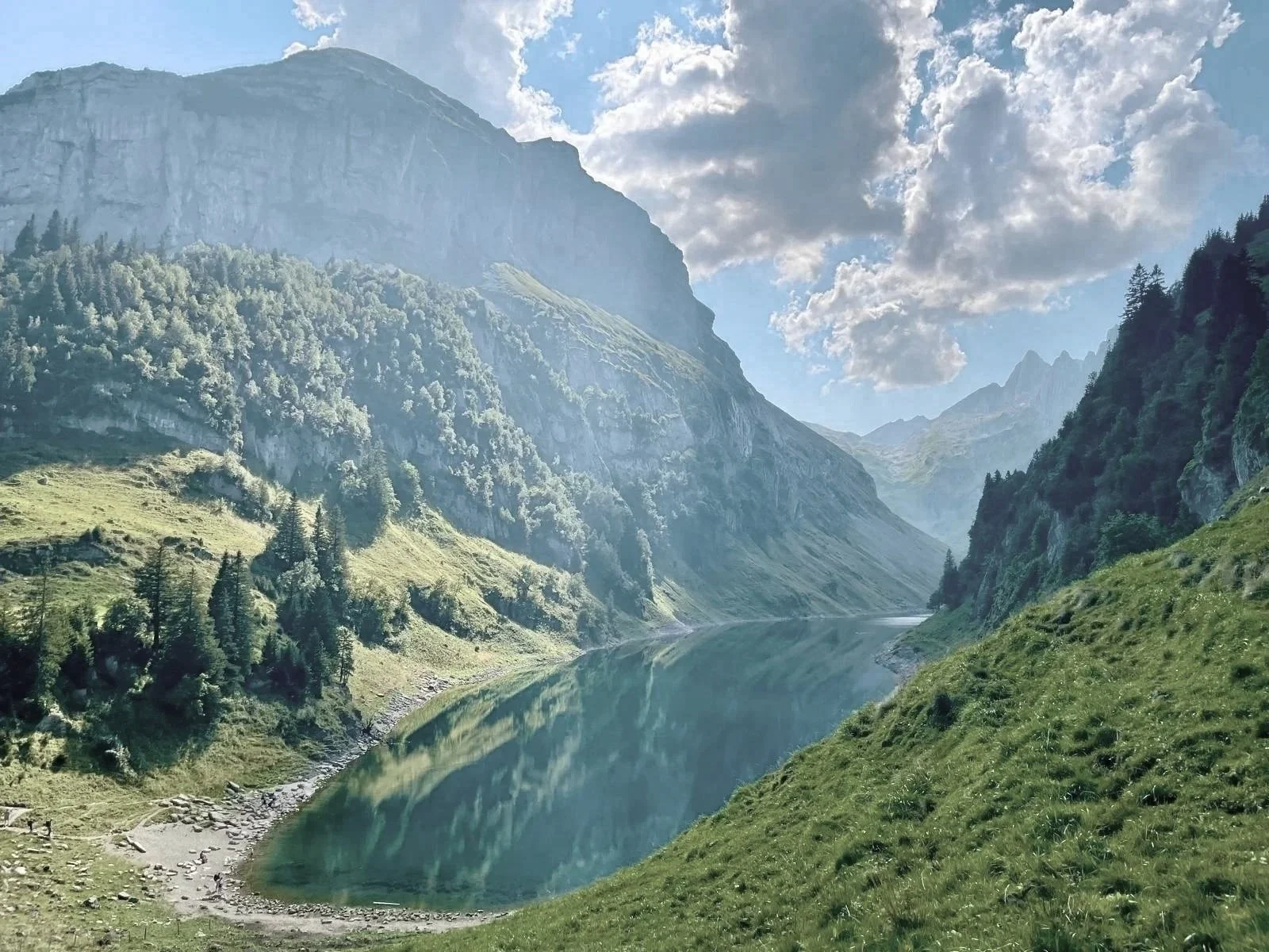 Berglandschaft mit einem ruhigen See, umgeben von grünen Hängen und hohen, bewaldeten Bergen unter einem bewölkten Himmel.