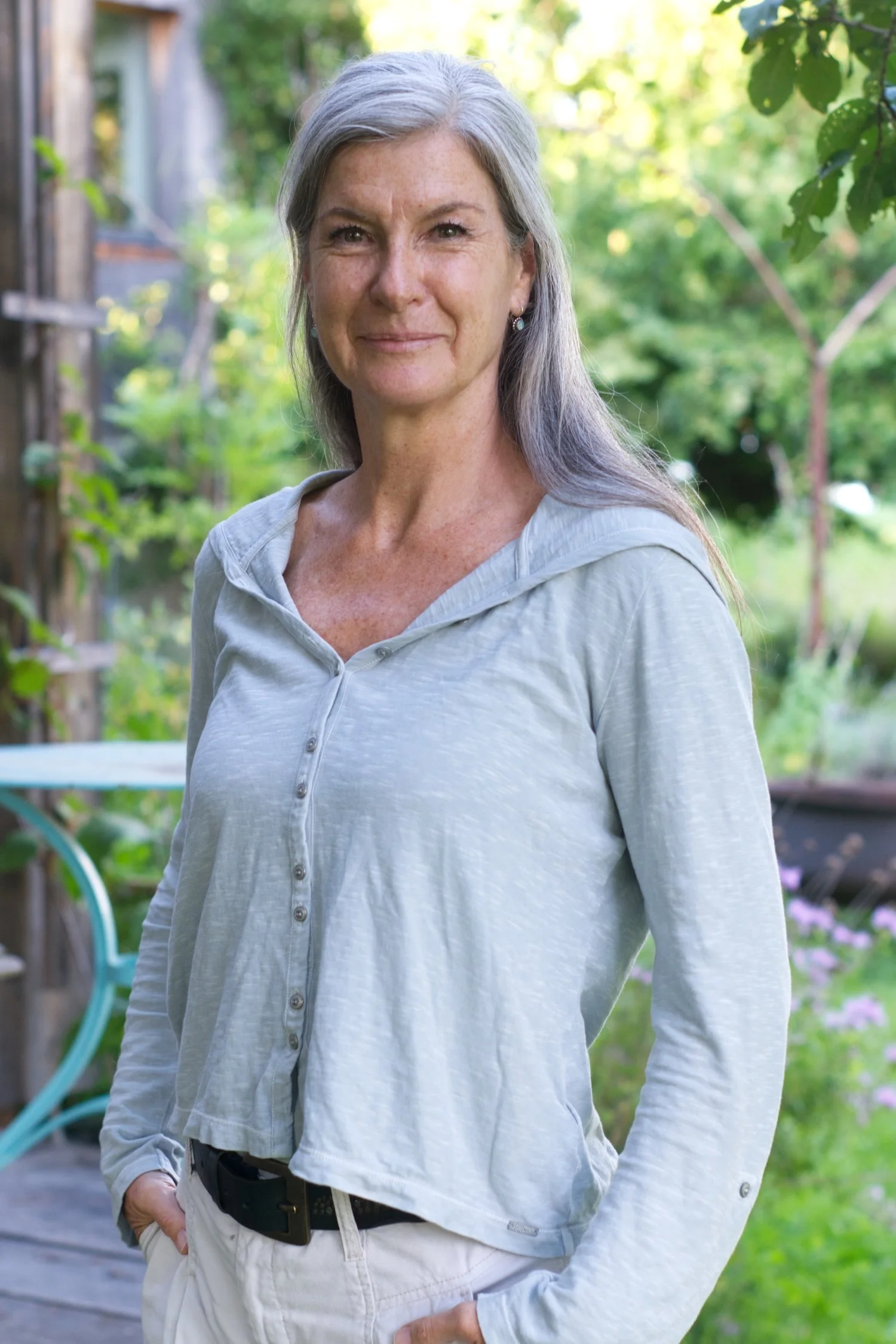 Eine lächelnde Frau mit grauem Haar in einer hellblauen Bluse, im Garten mit grünen Bäumen und bunten Blumen im Hintergrund.