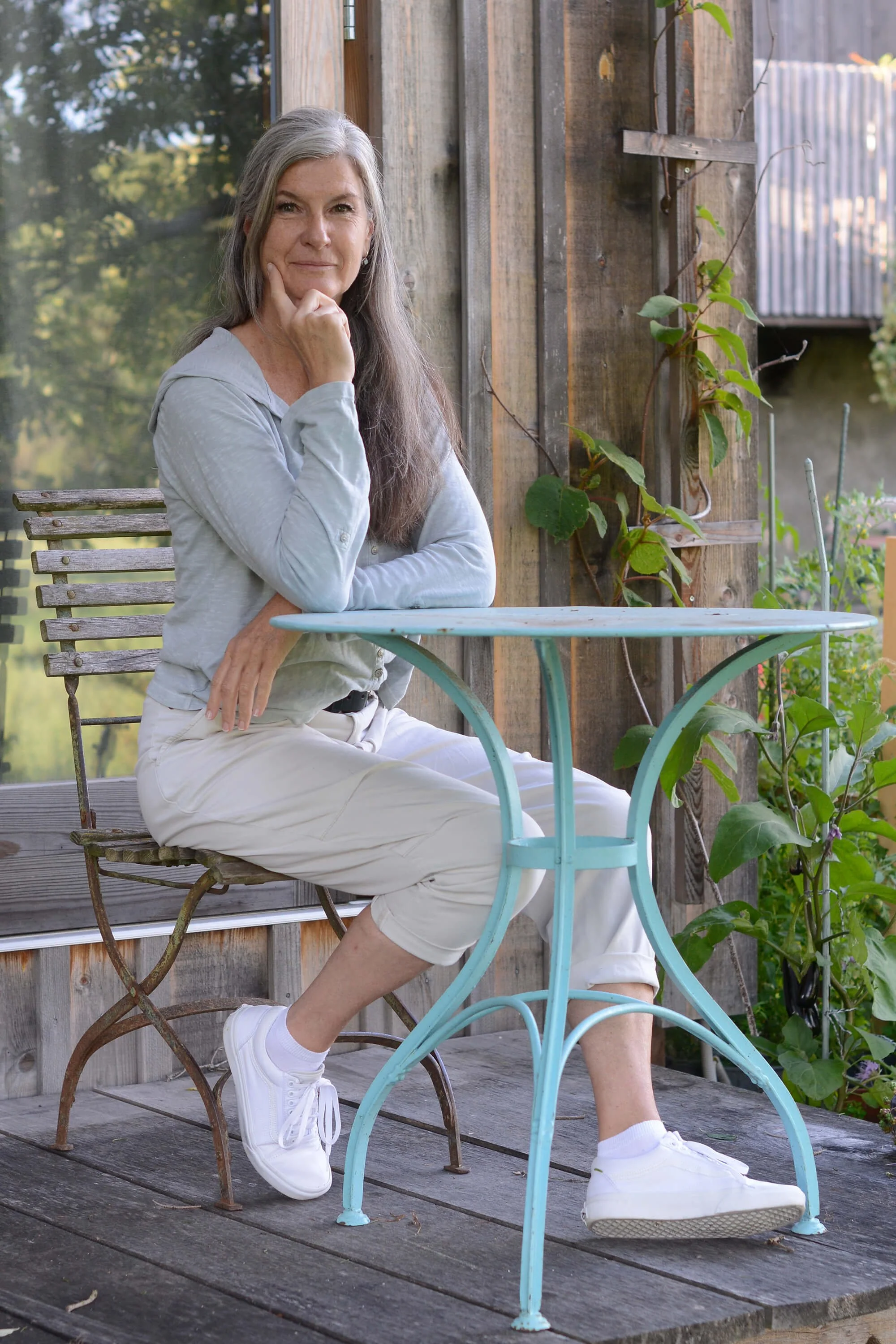 Eine Frau mit langen grauen Haaren sitzt auf einer Gartenbank auf einer Holzterrasse und blickt nach vorne, während sie die rechte Hand auf das Kinn stützt. Sie trägt eine hellblaue Bluse, weiße Hosen, weiße Turnschuhe und weiße Socken. Im Hintergrund sind grüne Pflanzen und ein Holzzaun sichtbar.