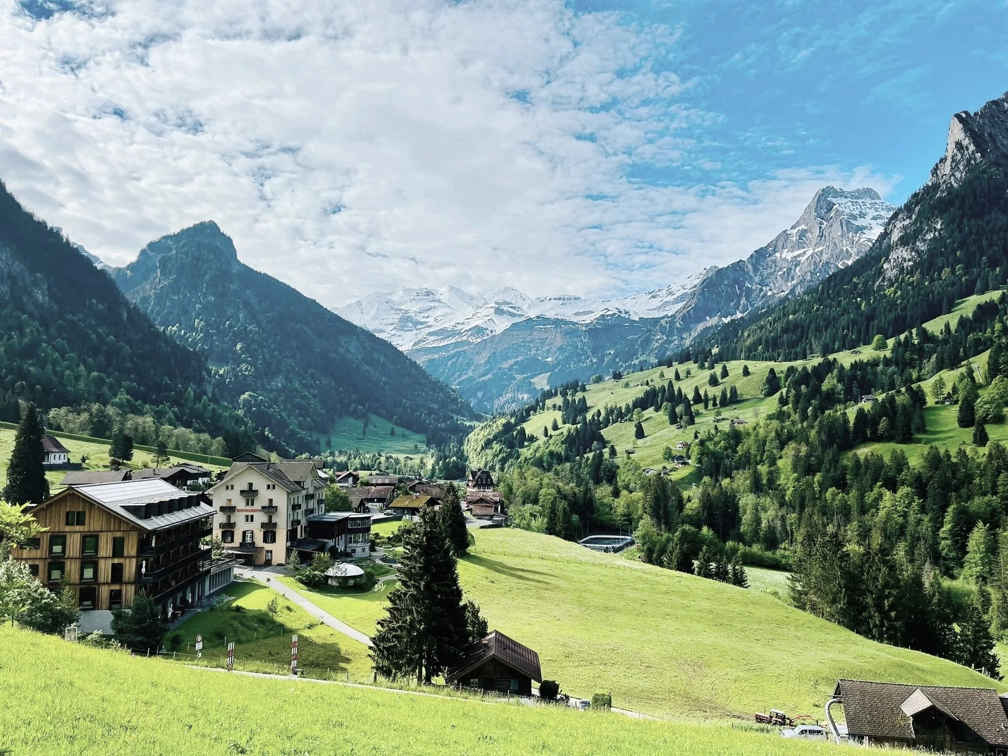 Ein malerisches Bergdorf in einer grünen Hügellandschaft mit Bergen im Hintergrund, einige Häuser im Vordergrund und bewölkter Himmel.