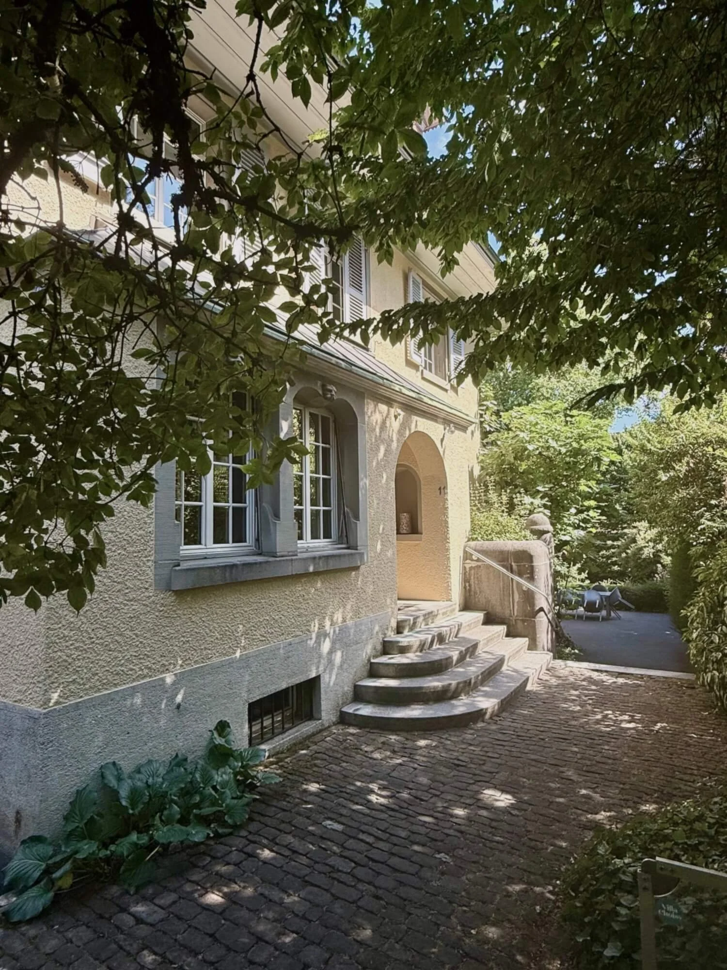Ein Haus mit gelber Fassade, weißen Fenstern und Steintreppen, umgeben von grünen Laubbäumen und einer gepflasterten Terrasse bei Sonnenlicht.