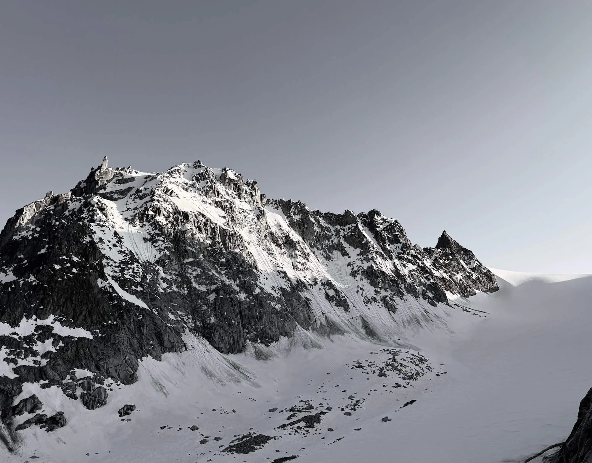 Gipfel eines verschneiten Berges mit steilen, felsigen Hängen und einem Gletscher im Vordergrund, bei bewölktem Himmel.