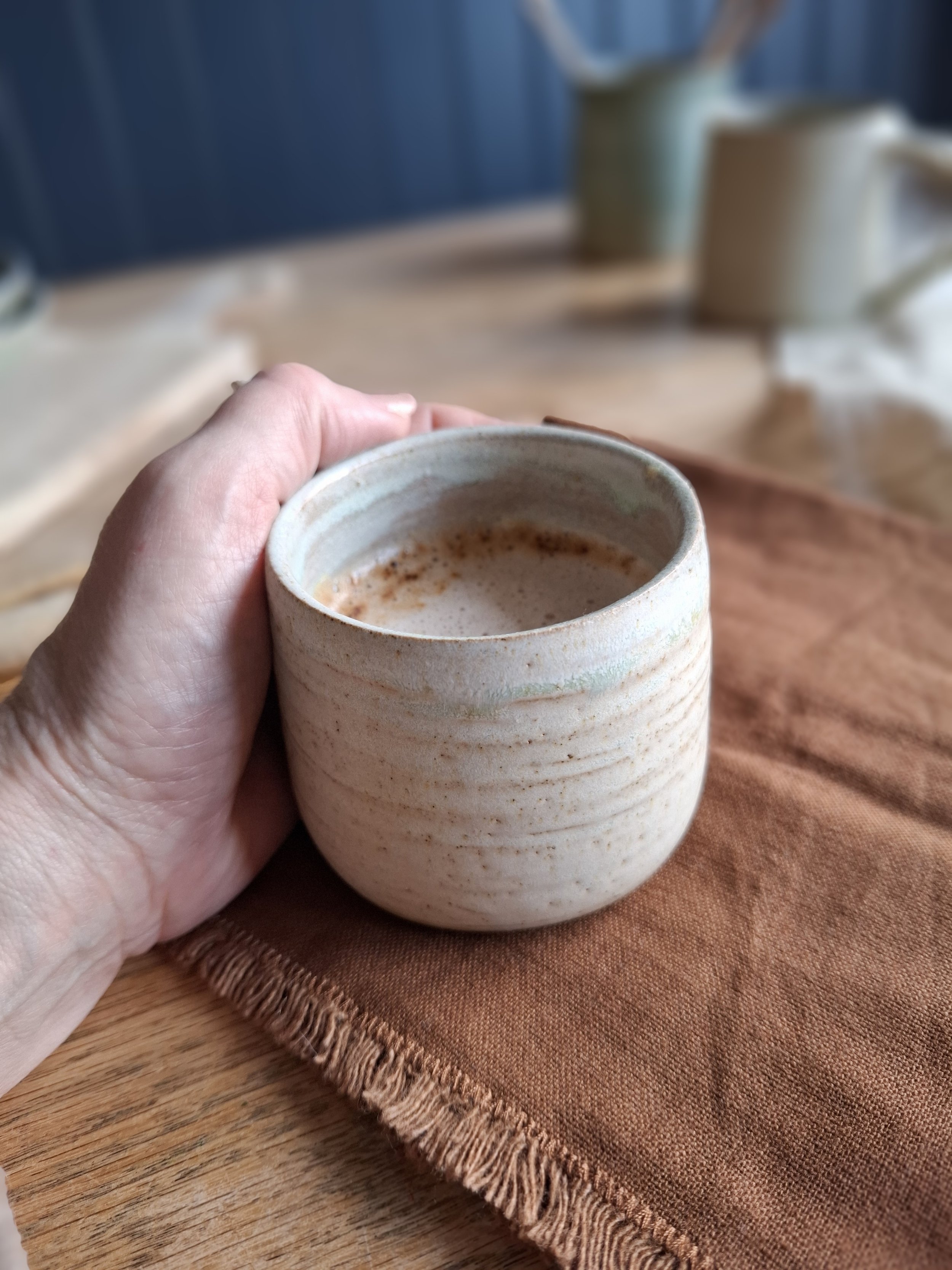 A hand holding a ceramic cup filled with a hot beverage, placed on a brown cloth on a wooden table.