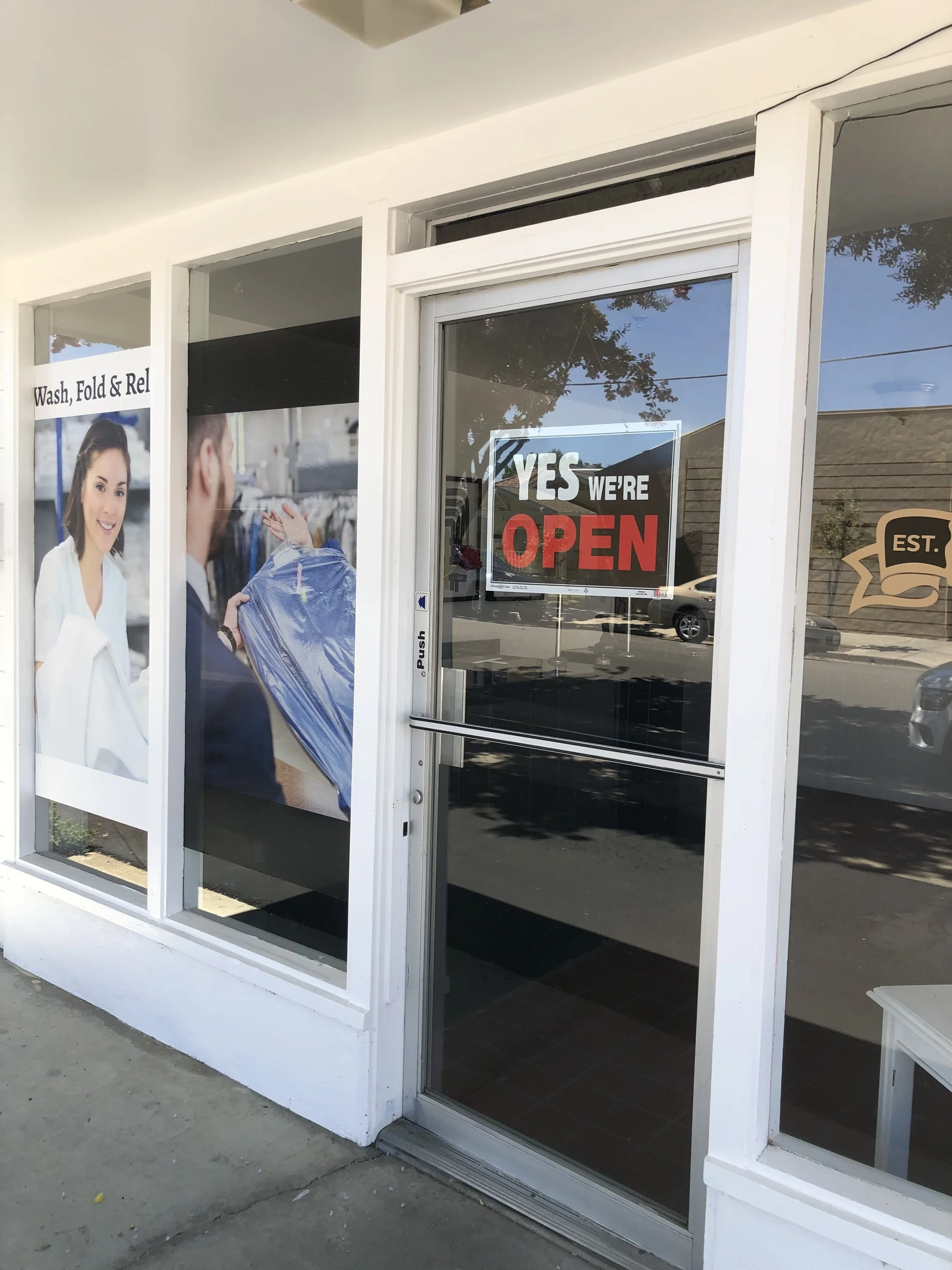 Storefront with large glass door and windows, sign on door says 'YES WE'RE OPEN', promotional poster in window shows a woman smiling in a white coat and a man holding a garment, sidewalk in front.
