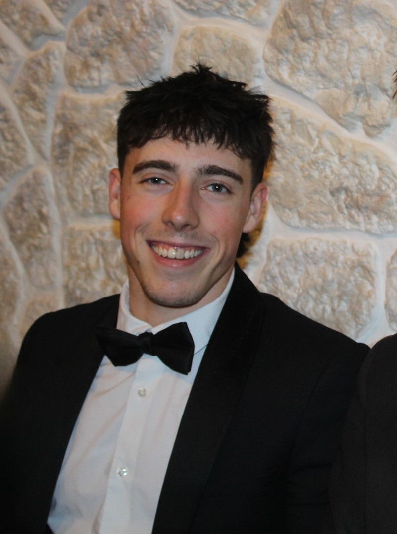 Young man dressed in a black tuxedo with a bow tie, smiling in front of a stone wall.