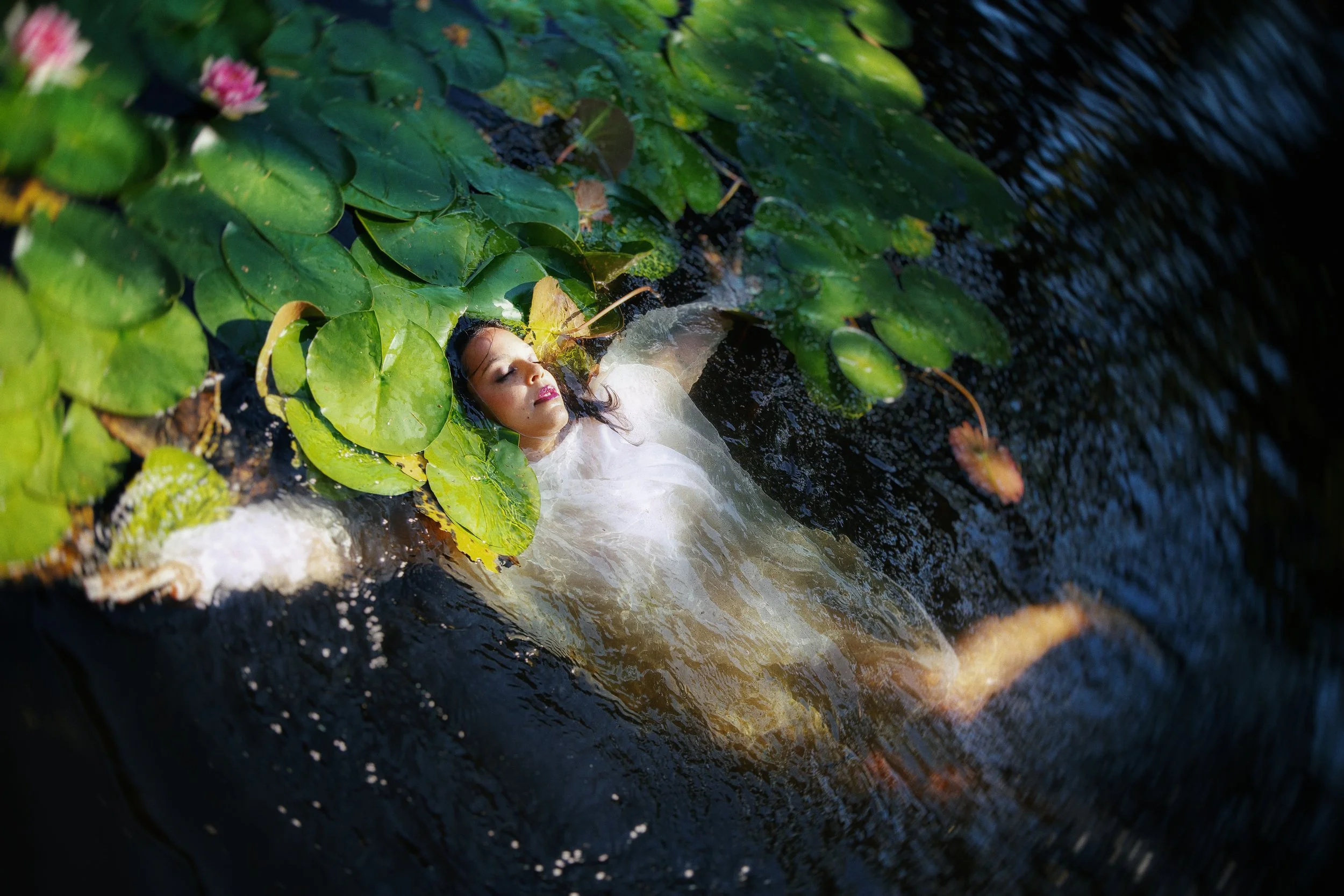 Frau mit weißen Kleid liegt im Wasser unter grünen Seerosenblättern in einem Fluss oder Teich, umgeben von dunklem Wasser.