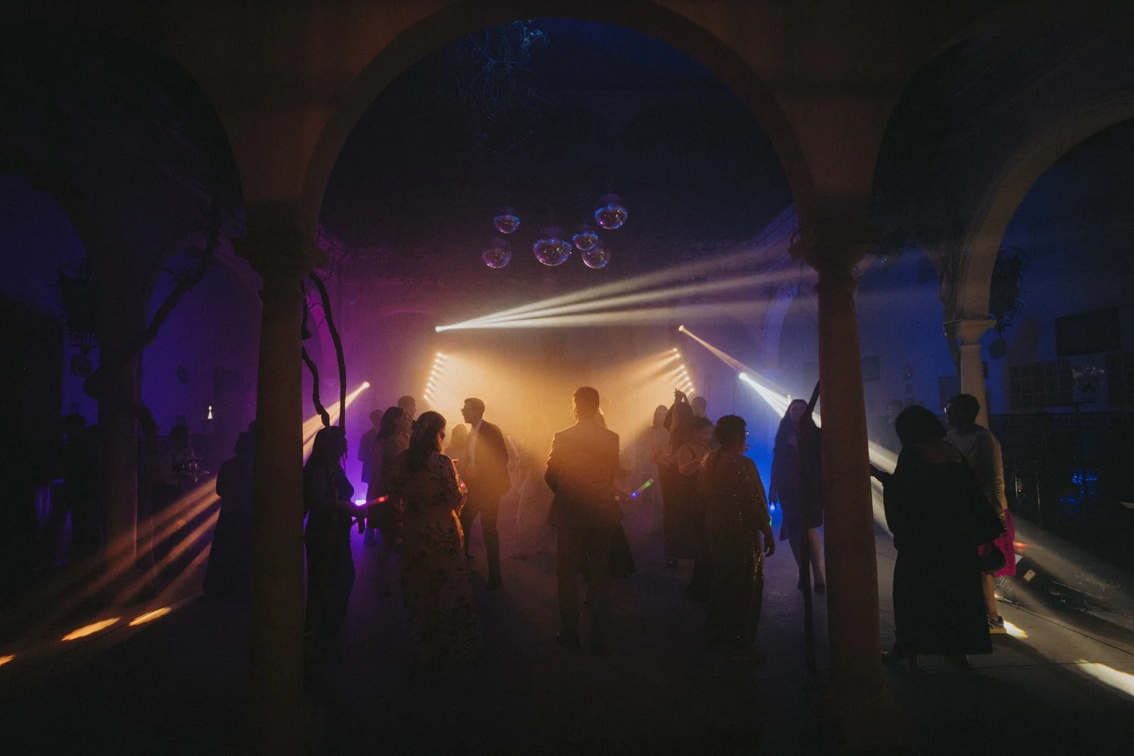 People dancing and socializing in a dimly lit nightclub or party with colorful lighting and disco balls overhead.