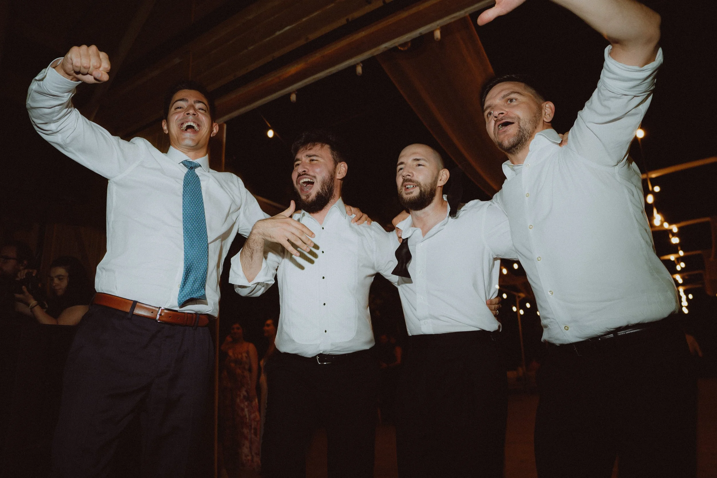 Four men in dress shirts and one with a tie, happily celebrating together with their arms around each other in a dimly lit indoor setting.