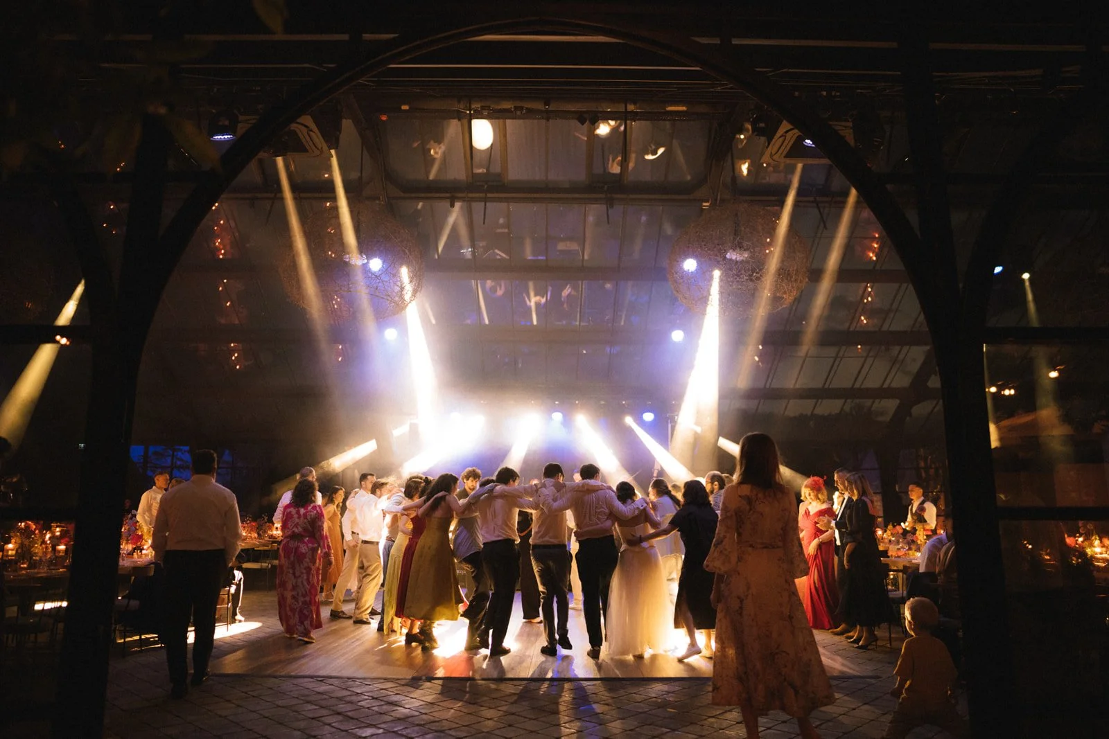 People dancing in a circle at a formal event with colorful hanging decorations and warm lighting.