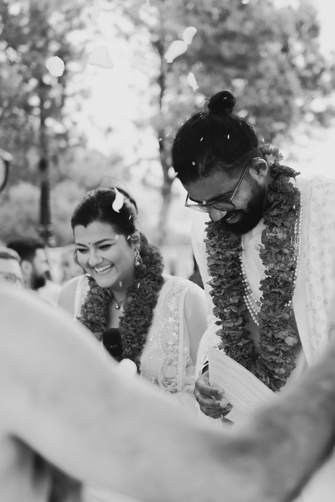 A couple dressed in traditional Indian attire, smiling and taking part in a wedding ceremony with garlands around their necks, outdoors with trees in the background.