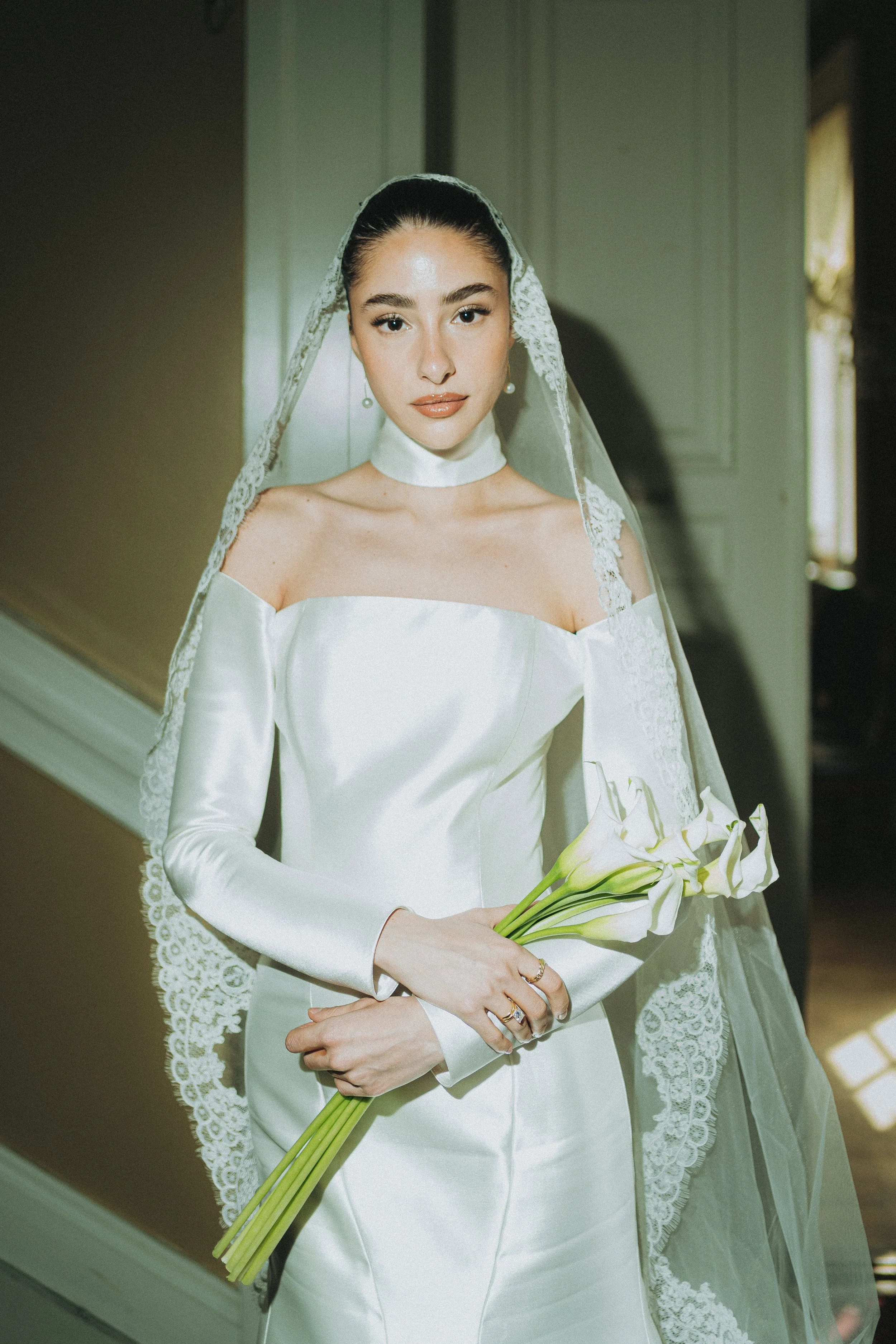 A young woman in a white wedding dress with lace detailing, holding a bouquet of white calla lilies, standing indoors with light coming through a window.