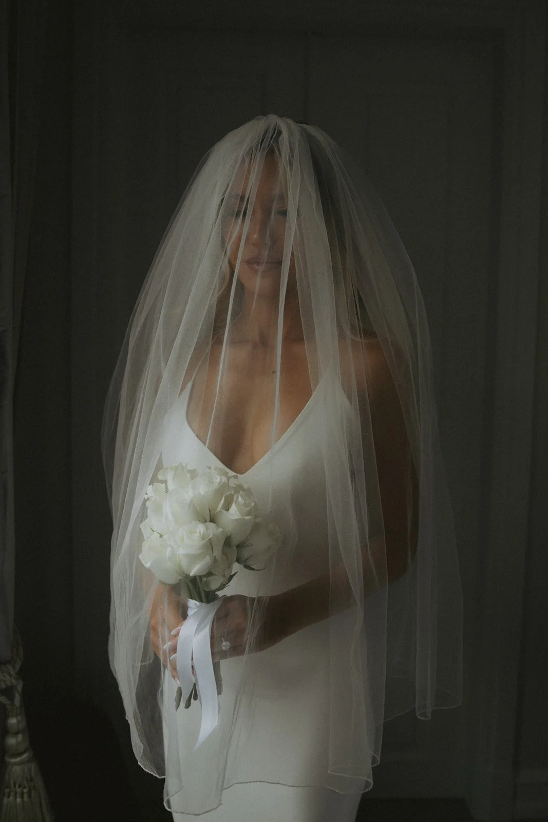A woman in a wedding dress holding a bouquet of white roses, wearing a veil, standing against a dark background.