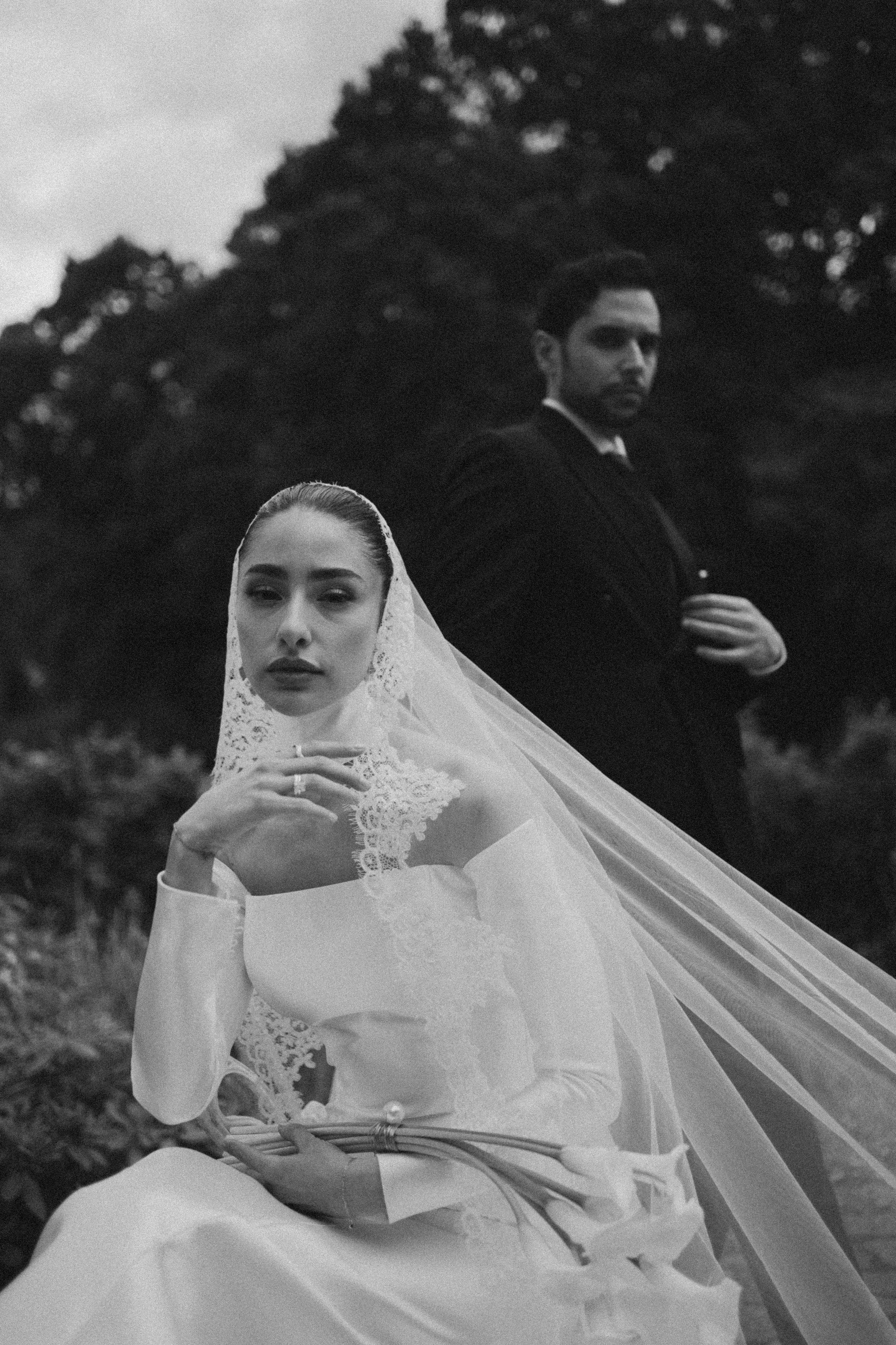 A black-and-white photo of a woman in a white lace dress and veil, posing outdoors with her hand resting on her chest, and a man in a suit standing behind her with his arms crossed, against a blurred background of trees.