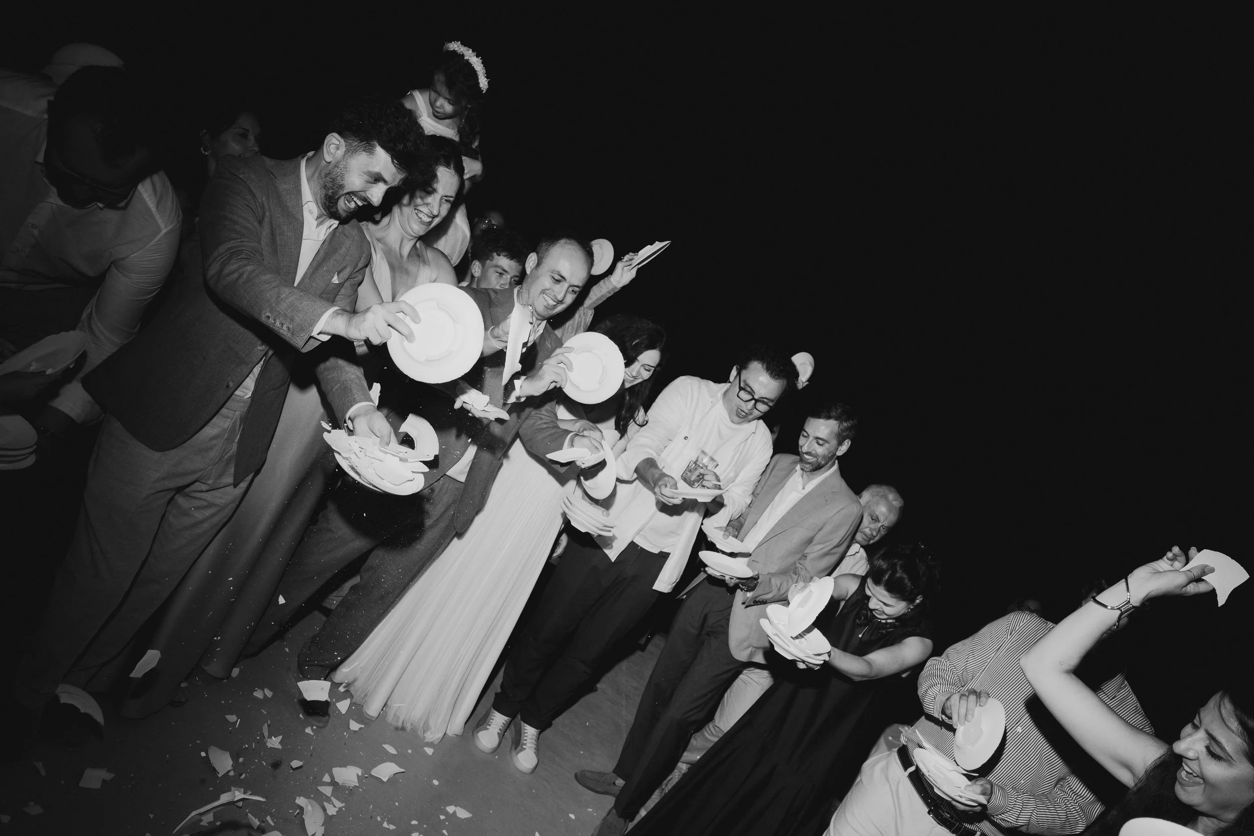 Group of people dressed in formal attire at a celebration, standing in a line, holding plates and cake, with some smiling and others looking down, against a dark background.